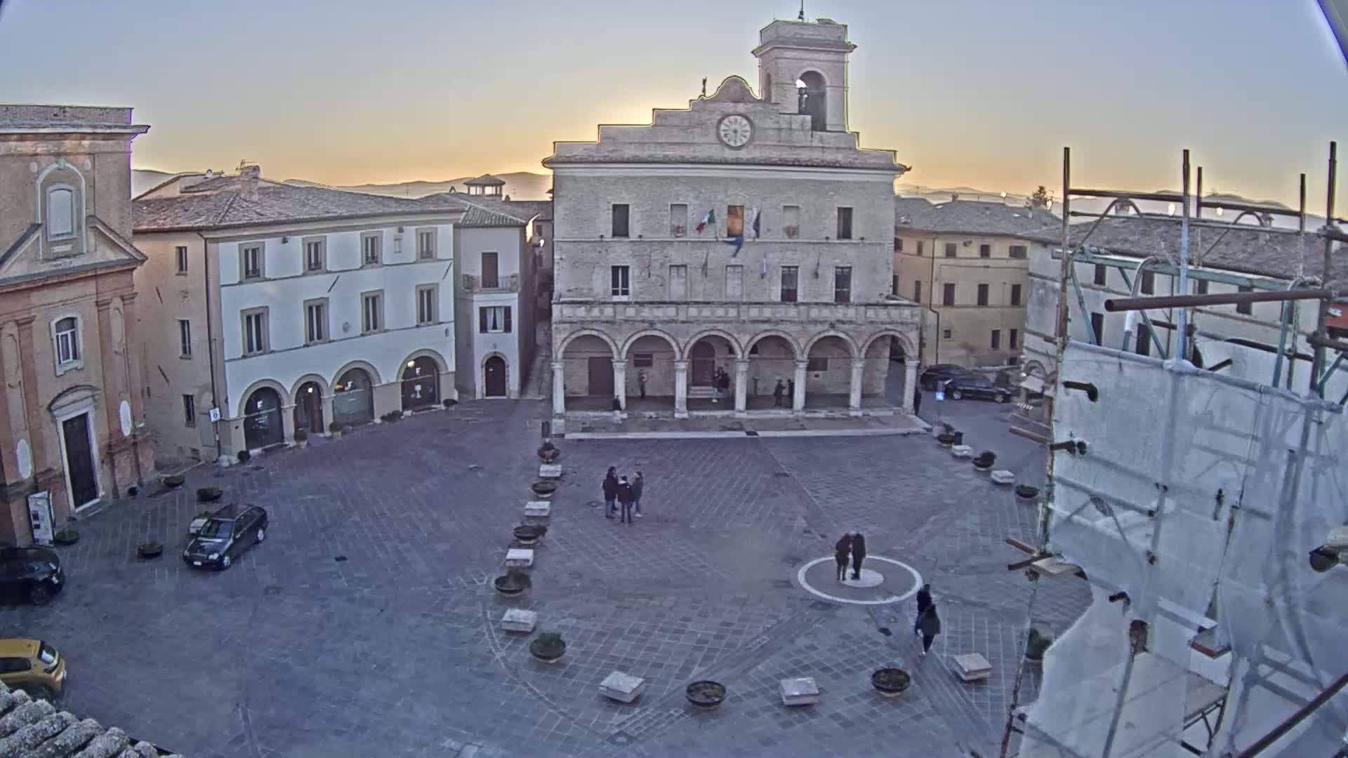 A large town square, under a sunrise sky, features a prominent stone building with arched entryways, surrounded by other buildings, with a few people and cars present.