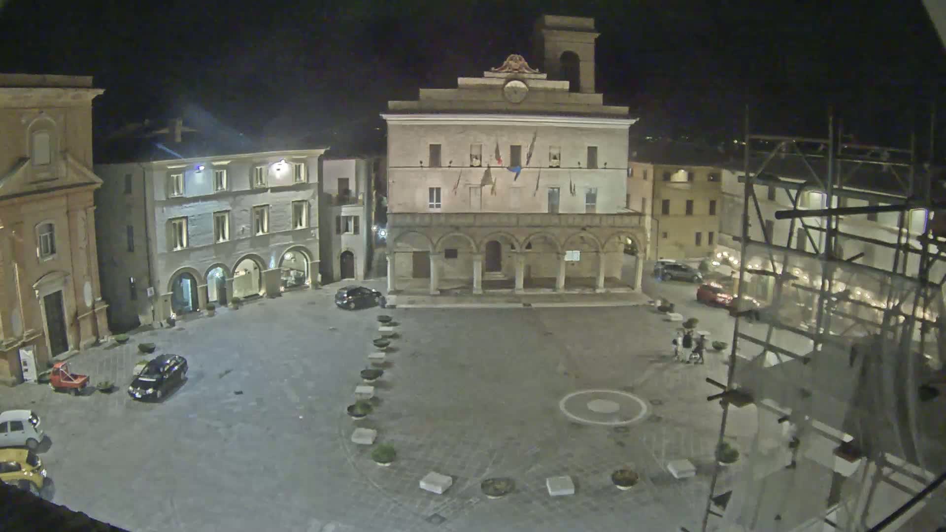 A town square at night, illuminated, with several buildings surrounding it, including a large building with arched entryways, and scaffolding in the bottom right.