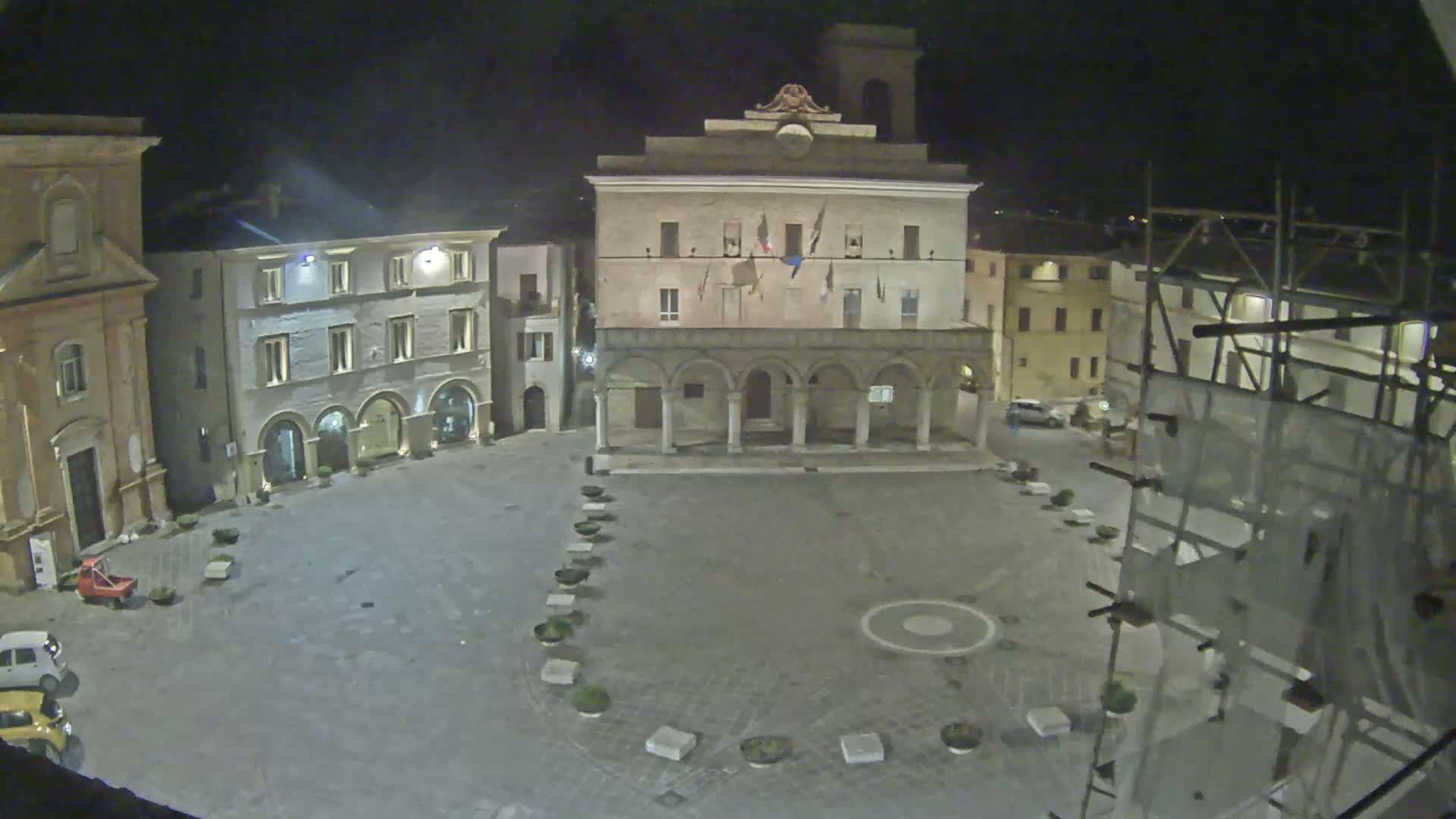 A nighttime view of an empty town square with several buildings surrounding it, including a large building with arches and a clock tower.