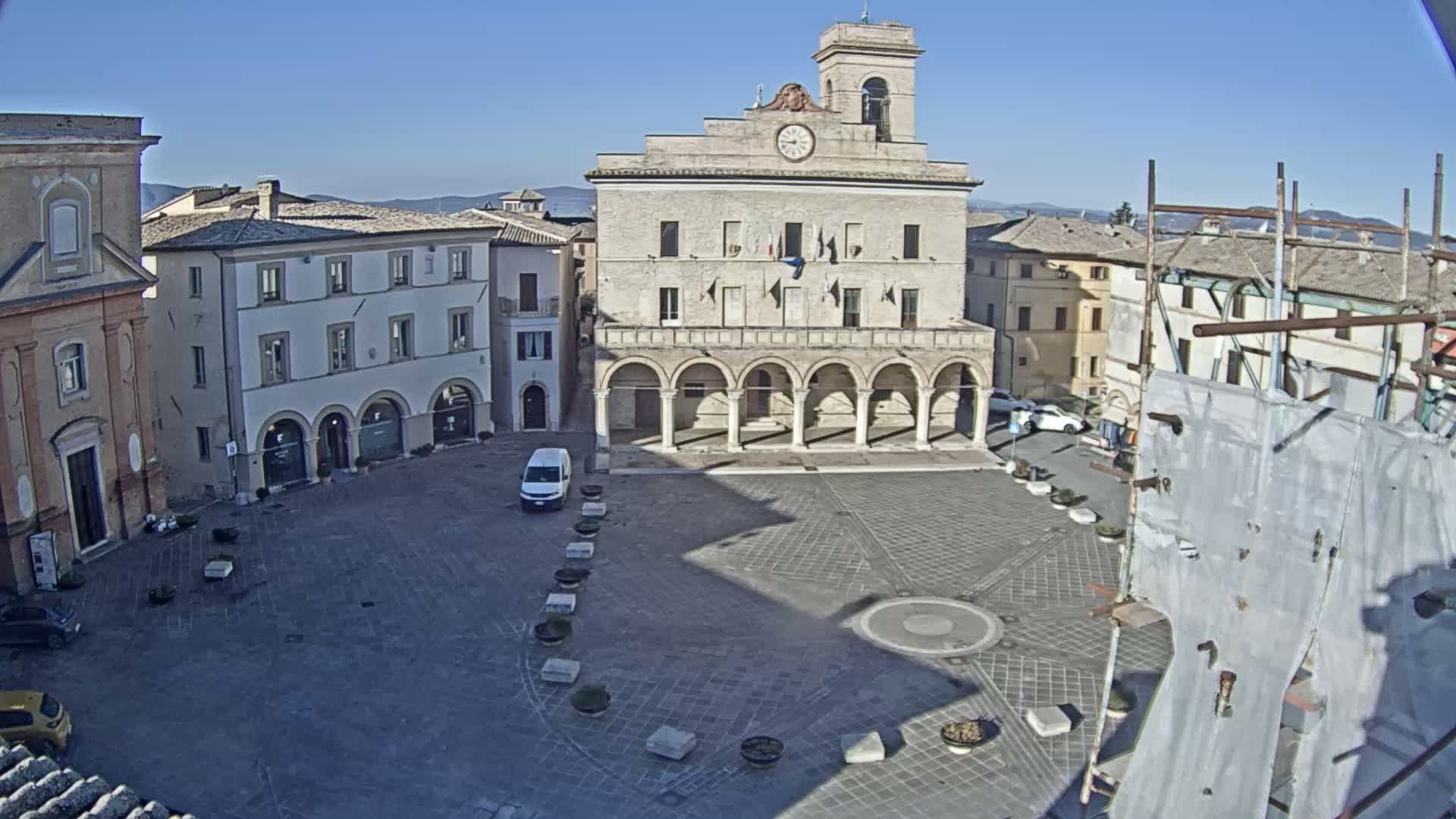 A town square with a large stone building featuring a clock tower and arched colonnade, under a clear blue sky.