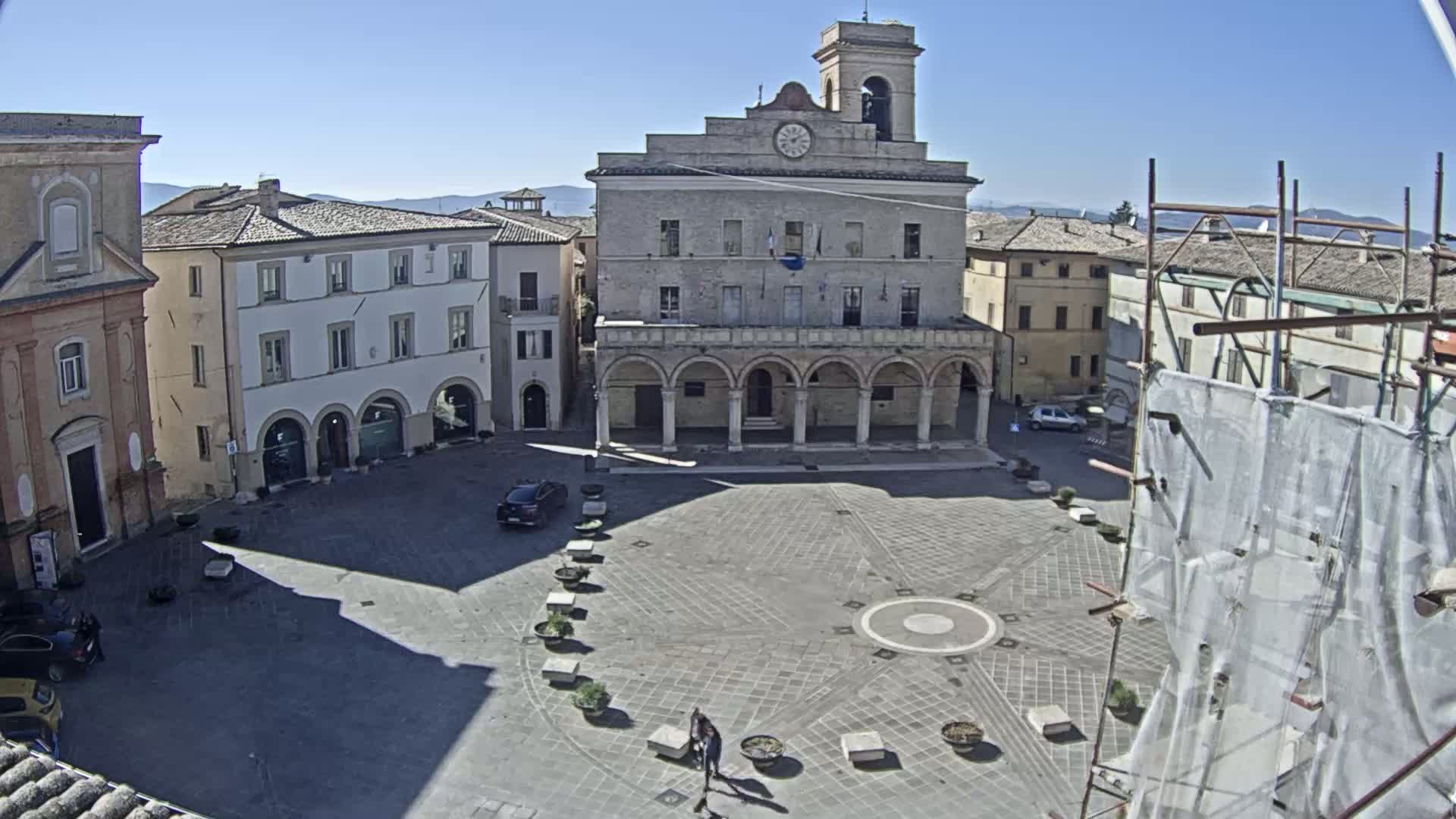 A large, stone town square with a clock tower and several buildings surrounding it on a sunny day.