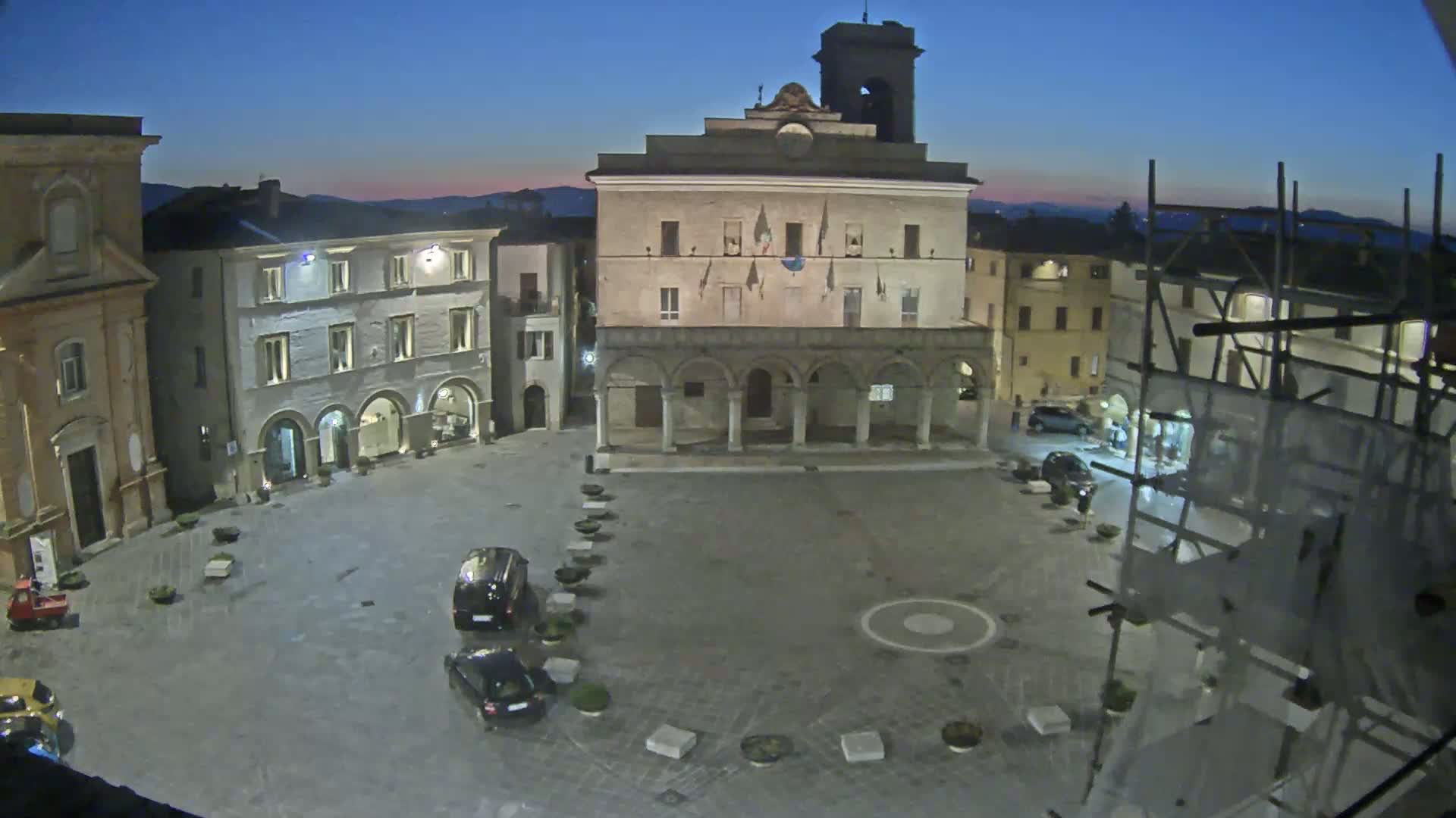 A town square at twilight, with several cars parked and buildings surrounding it, under a clear, dark blue sky.