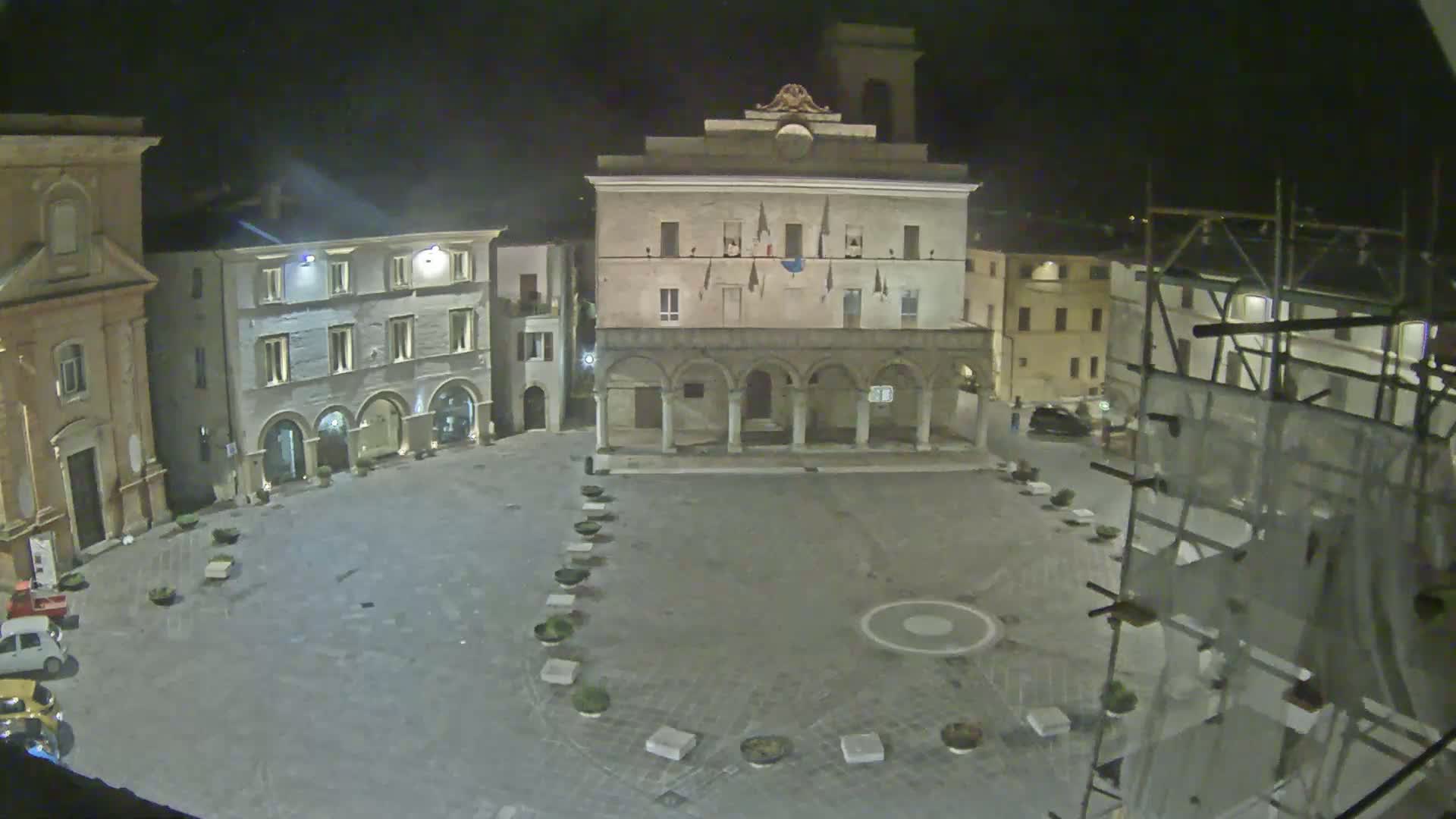 A nighttime, high-angle view of an empty town square with several historic buildings surrounding it, including one with a colonnade, and scaffolding on a building to the right.