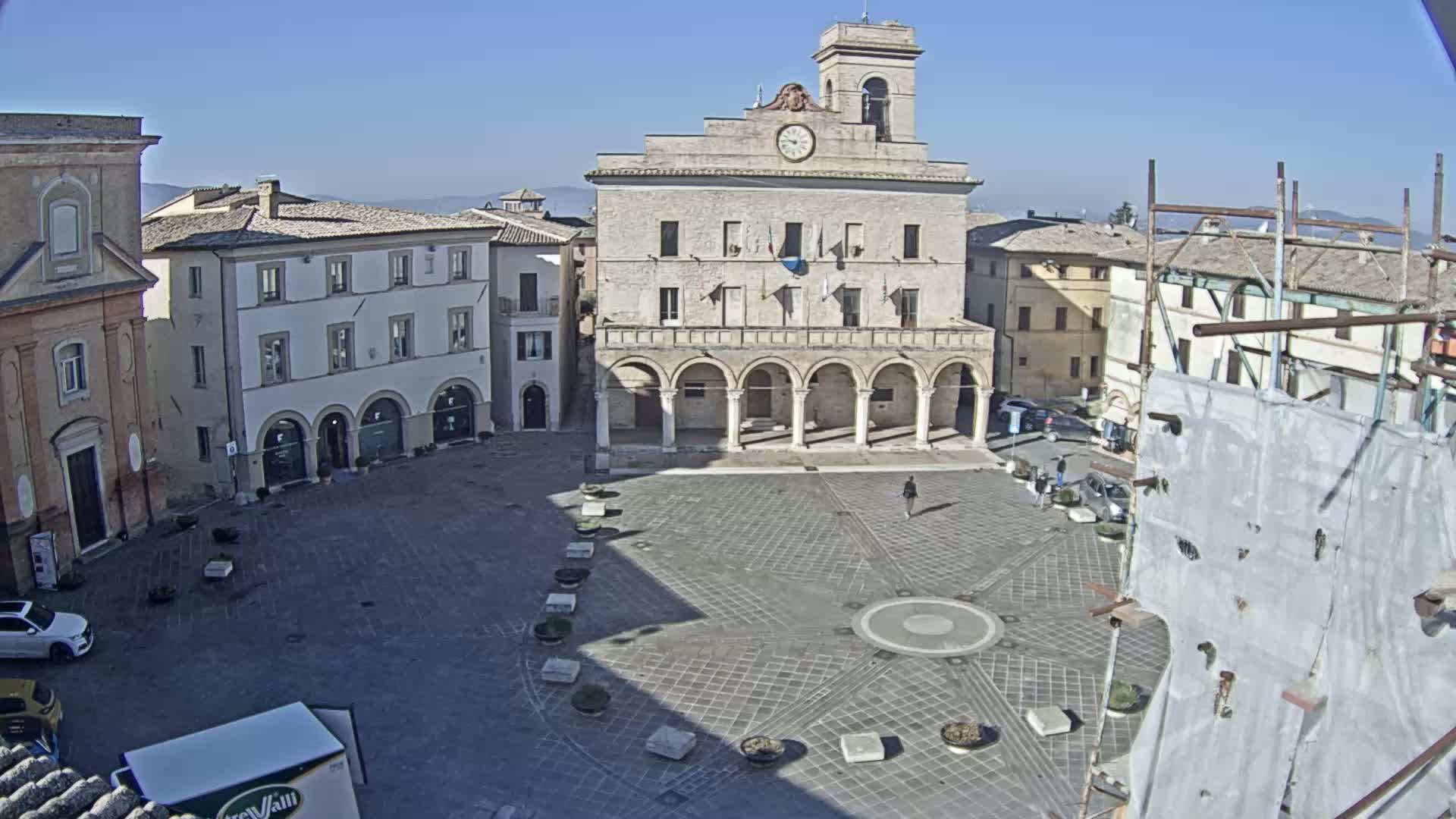 A mostly empty town square with a large, light-colored building featuring a clock tower and arched colonnade is seen under a clear, sunny sky.