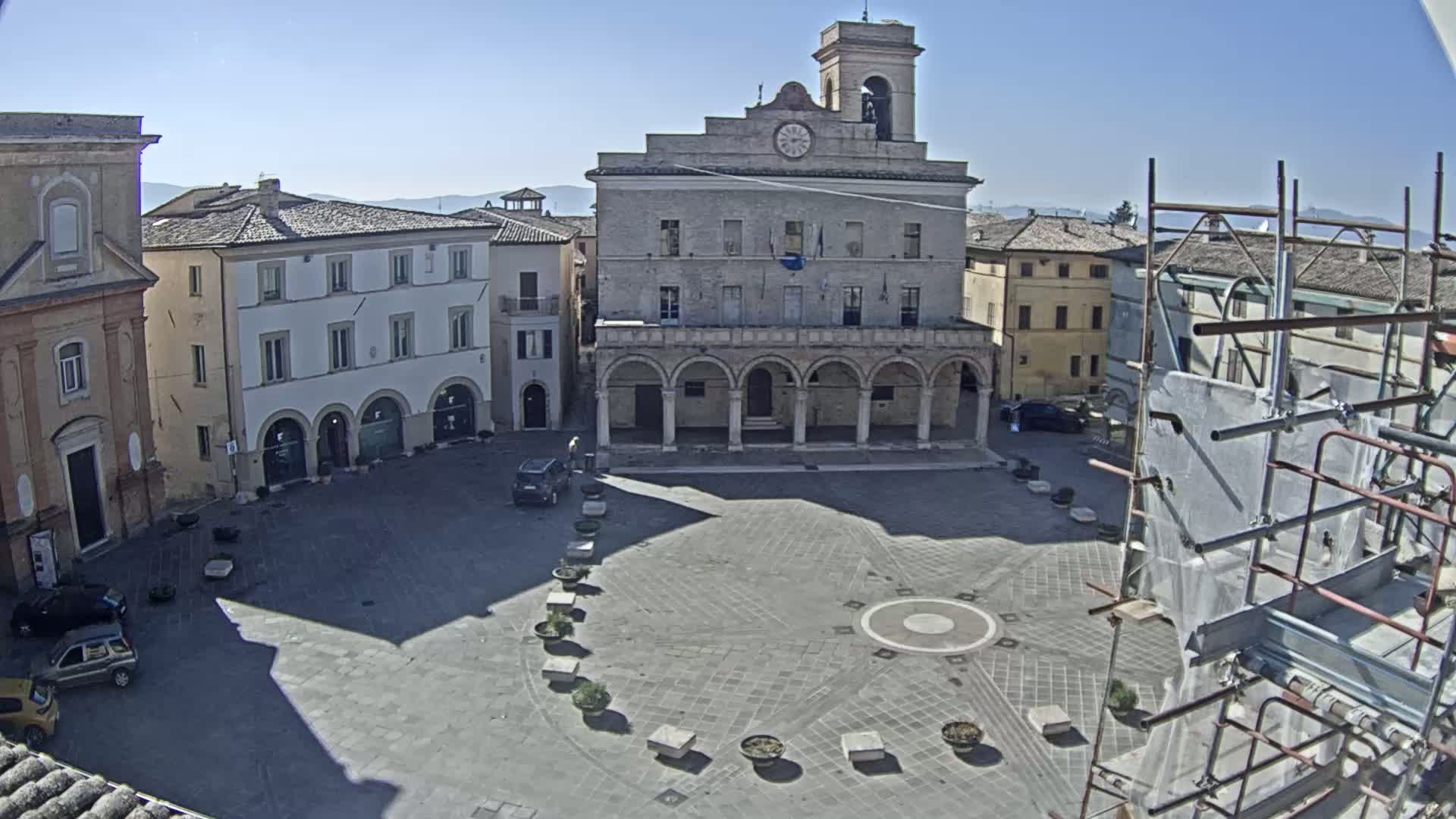 A sunny day reveals a town square centered on a large, classical-style building with arched colonnades, surrounded by other buildings and a few parked cars, with scaffolding partially obscuring the view in the lower right corner.