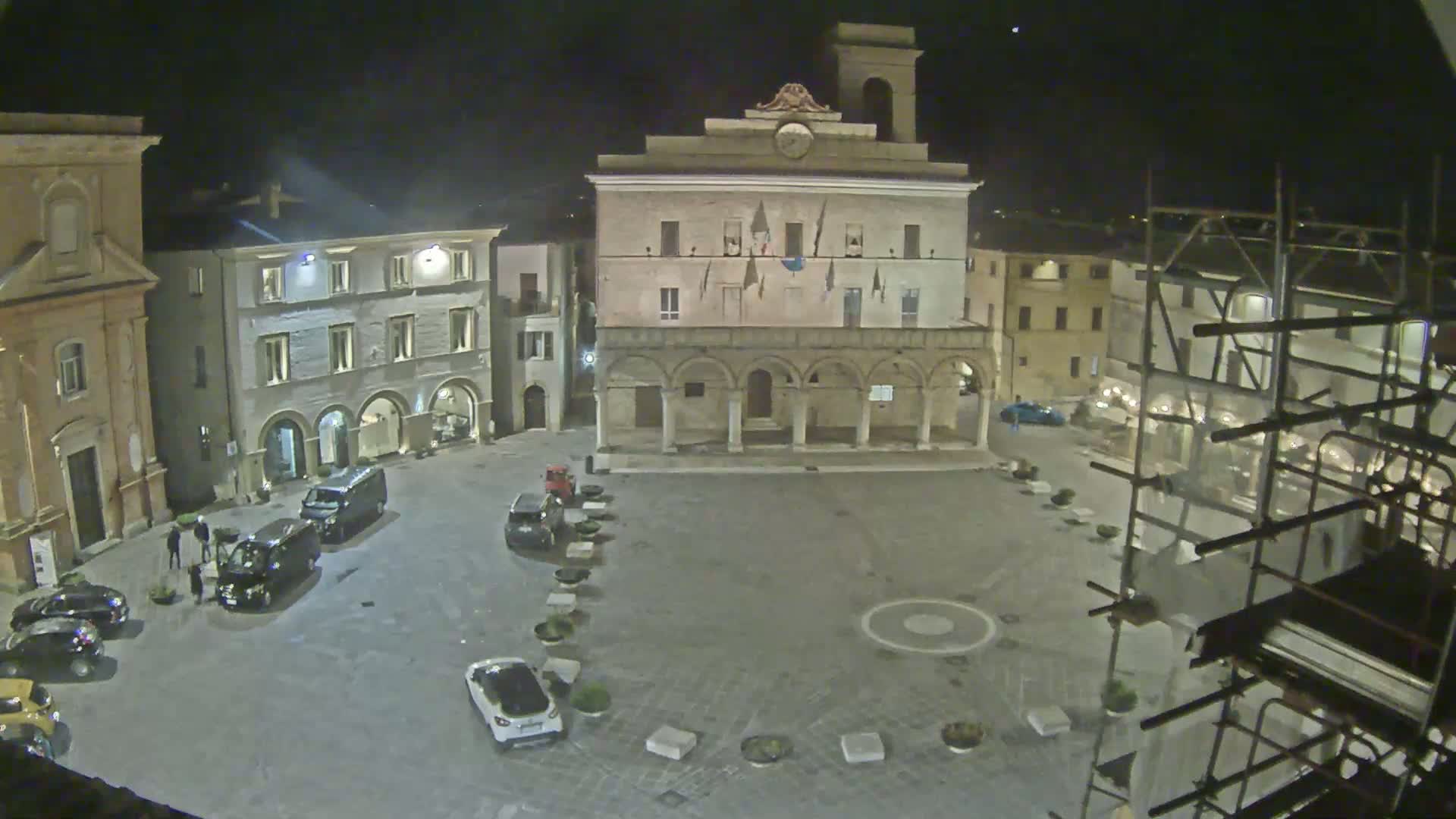 A nighttime view of a town square with several cars parked, buildings surrounding it, and scaffolding on one side.