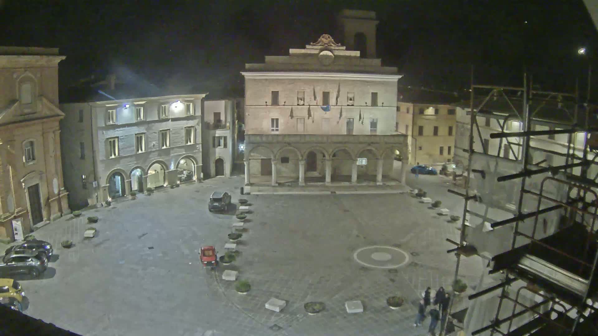 A nighttime view of a town square with several buildings, a few cars parked, and scaffolding on one building.