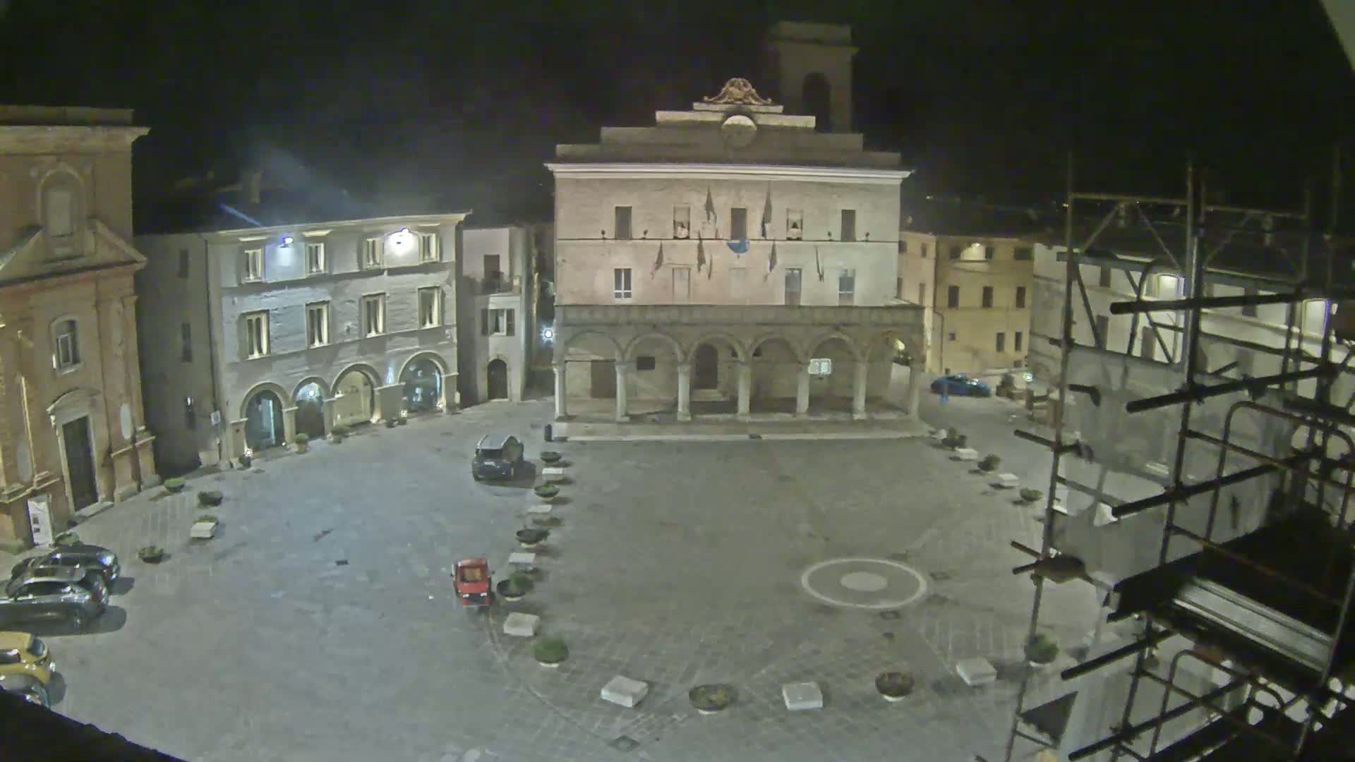 A nighttime view of a town square, featuring several buildings, parked cars, and scaffolding on one side, under a dark sky.