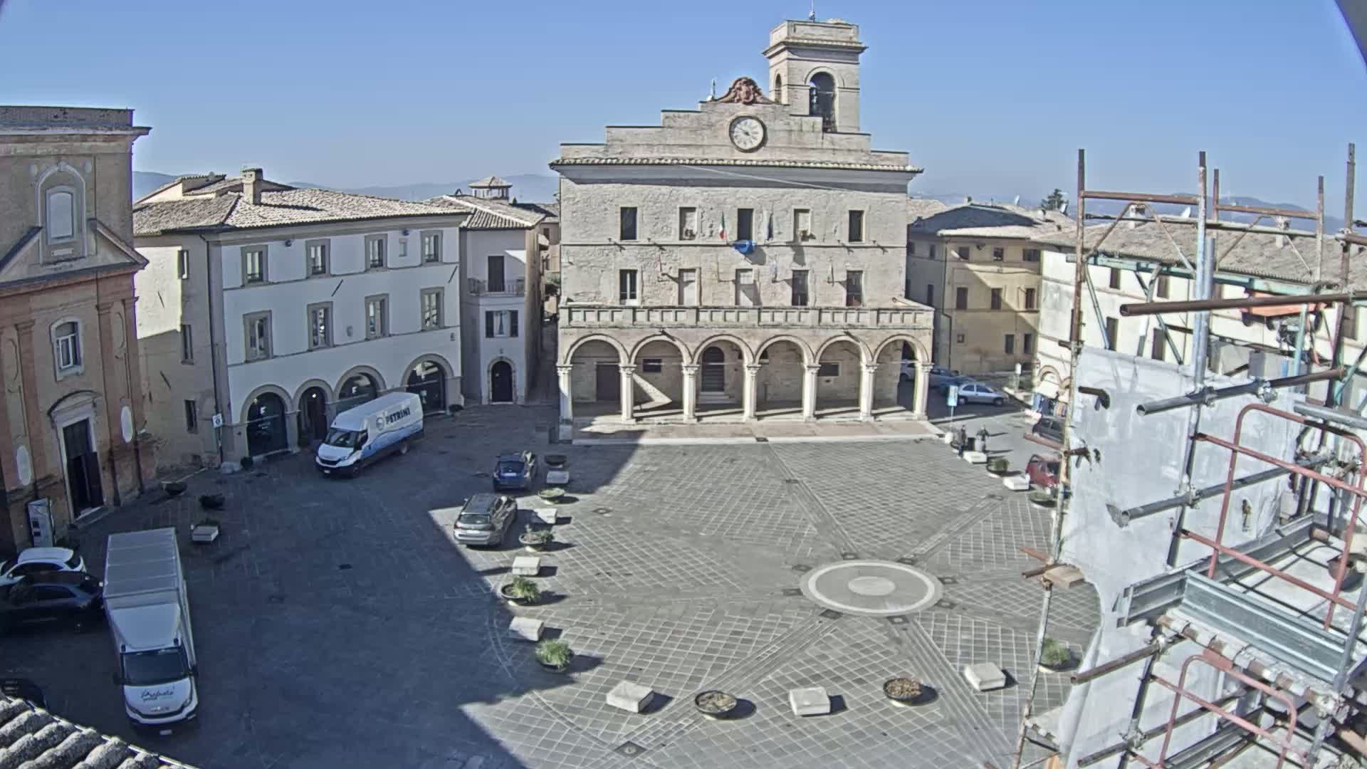 A large town square, under sunny skies, is centered on a classical-style building with a clock tower, surrounded by other buildings and a few parked cars.