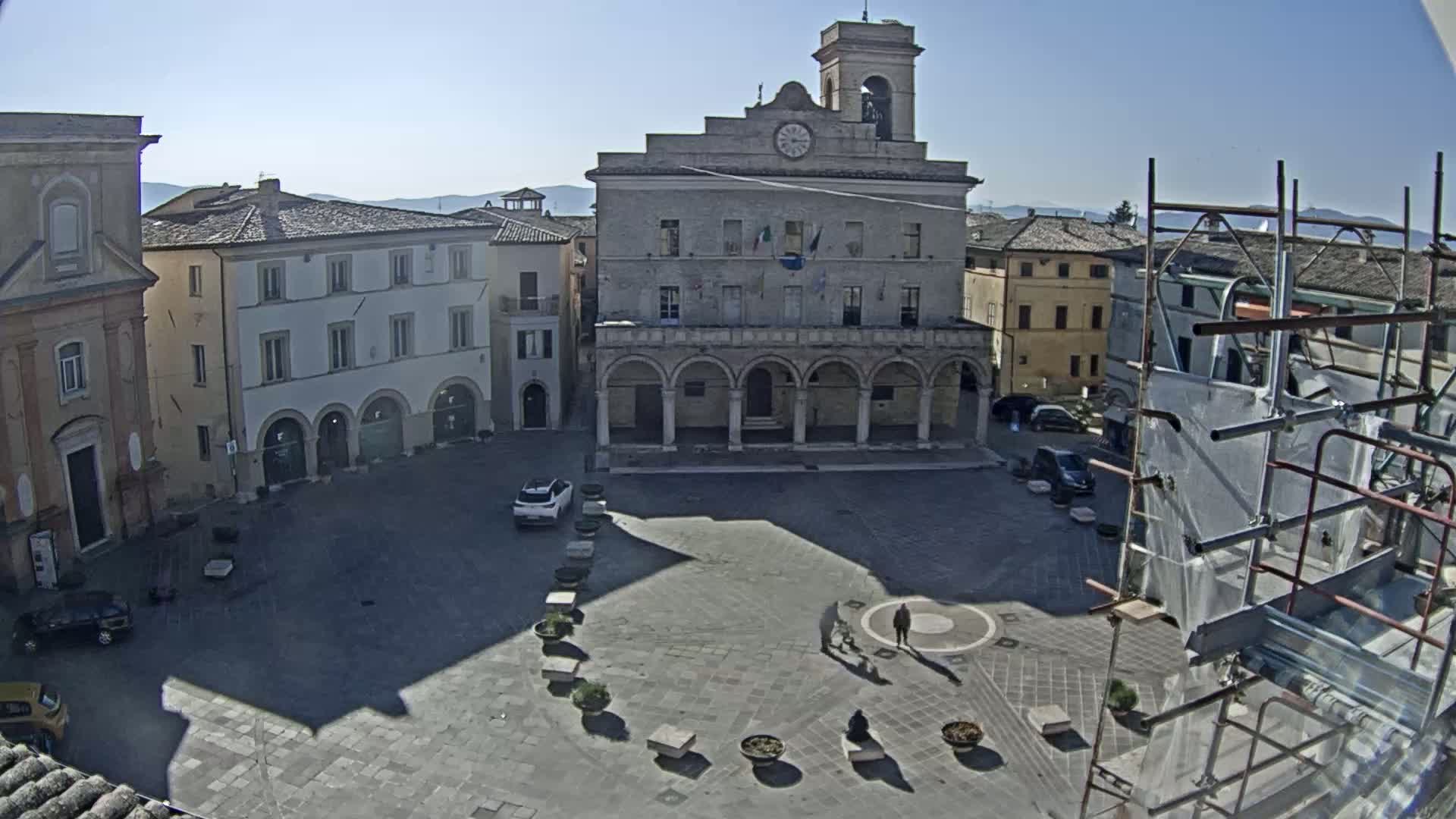 A mostly empty town square, under a bright sunny sky, is centered around a large building with arched lower windows and a clock tower.