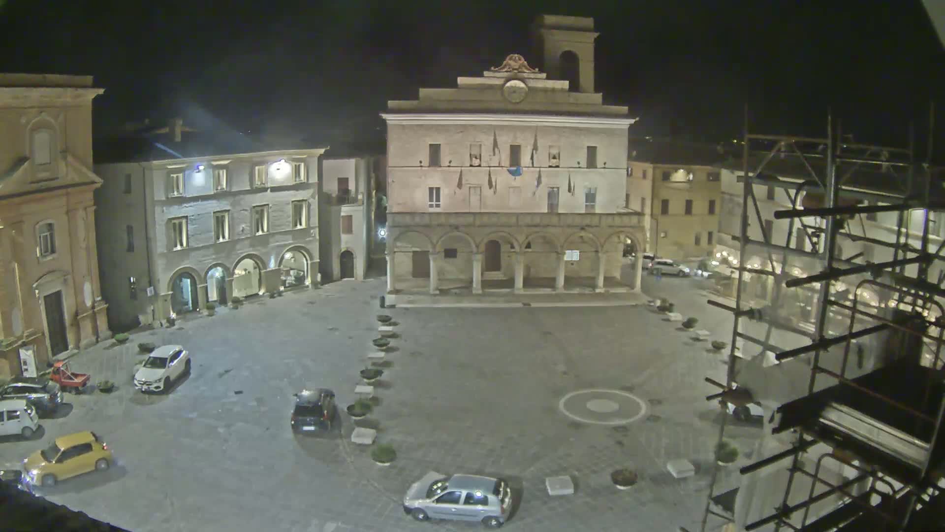 A nighttime view of a town square with several cars parked, buildings surrounding it, and scaffolding on one side.