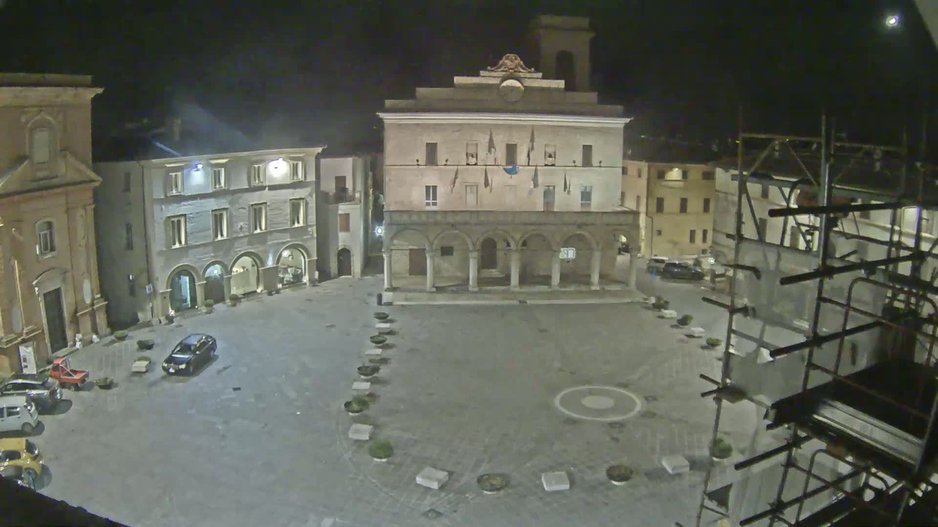 A nighttime view of a town square, featuring several buildings, a few parked cars, and scaffolding on one side, under a dark, clear sky.