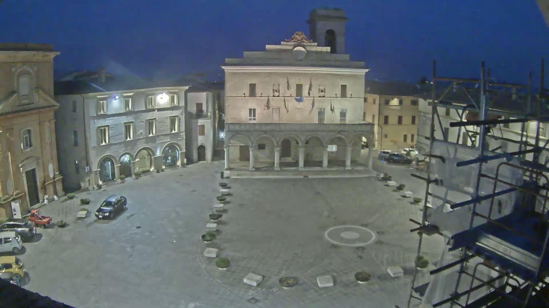 A mostly empty town square at night is surrounded by historic buildings, one of which is under construction.