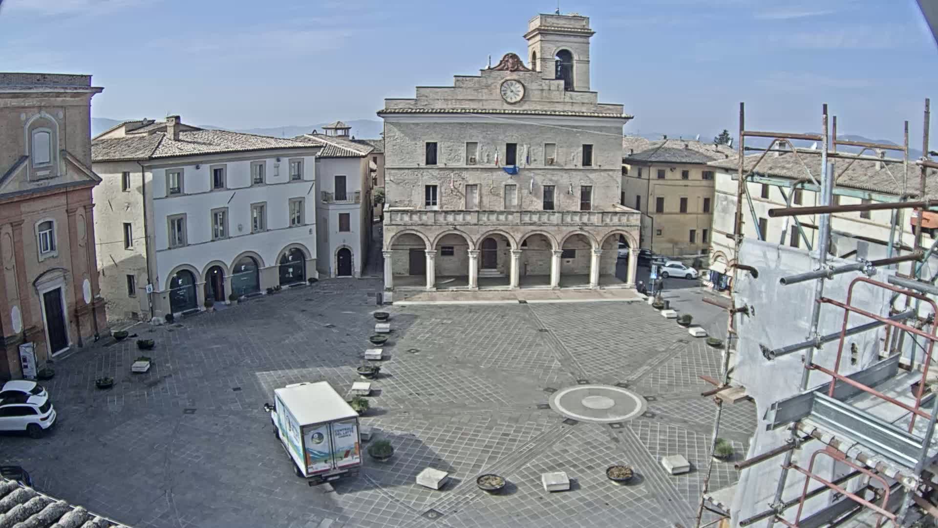 A large town square with a stone building featuring arched entryways, surrounded by other buildings, under a clear sky.