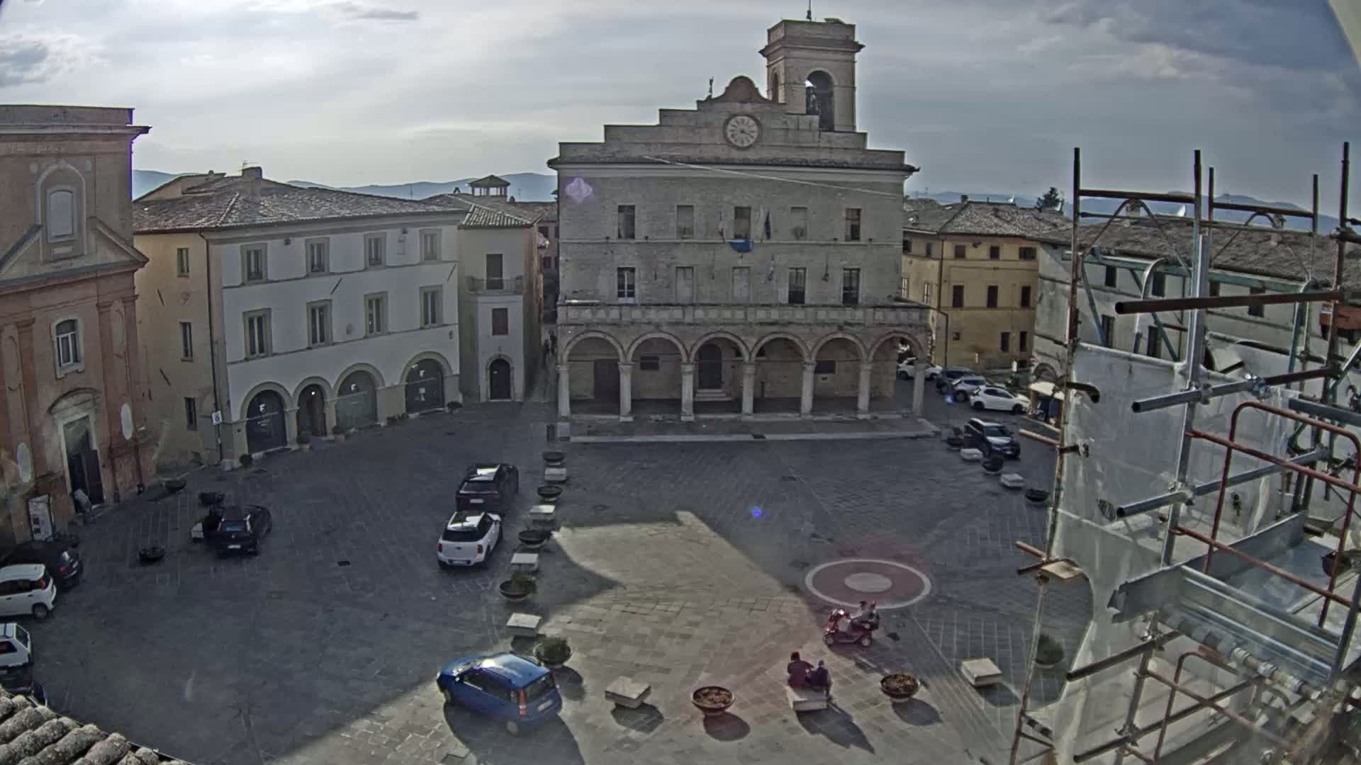 A town square, under a partly cloudy sky, is surrounded by historic buildings, with several cars parked and a few people sitting on benches.