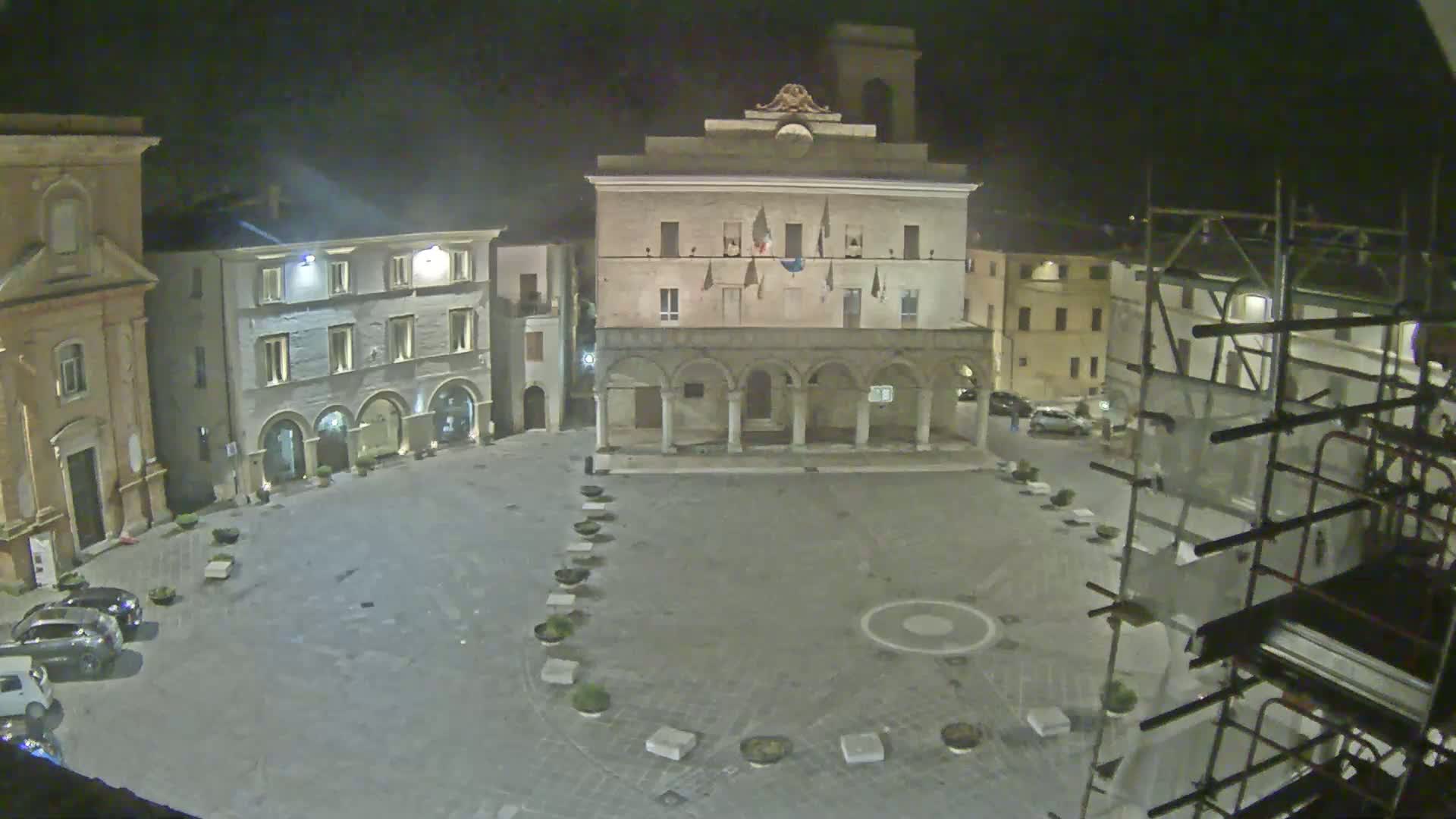 A nighttime view of a town square with a large building featuring arched entryways, surrounded by other buildings, some with scaffolding, under dark, clear skies.