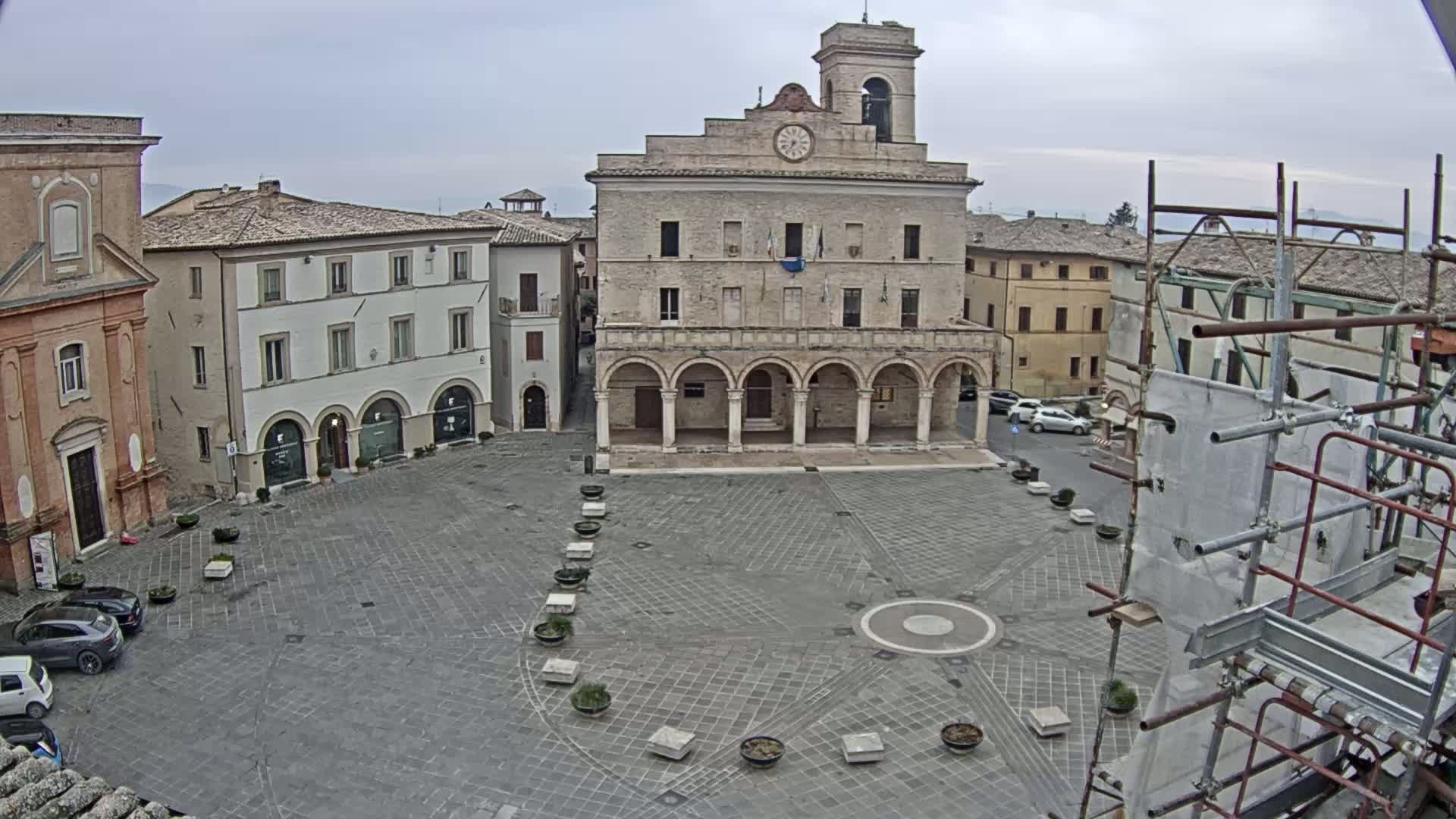A town square with a large stone building, several smaller buildings, and a few cars, under an overcast sky.