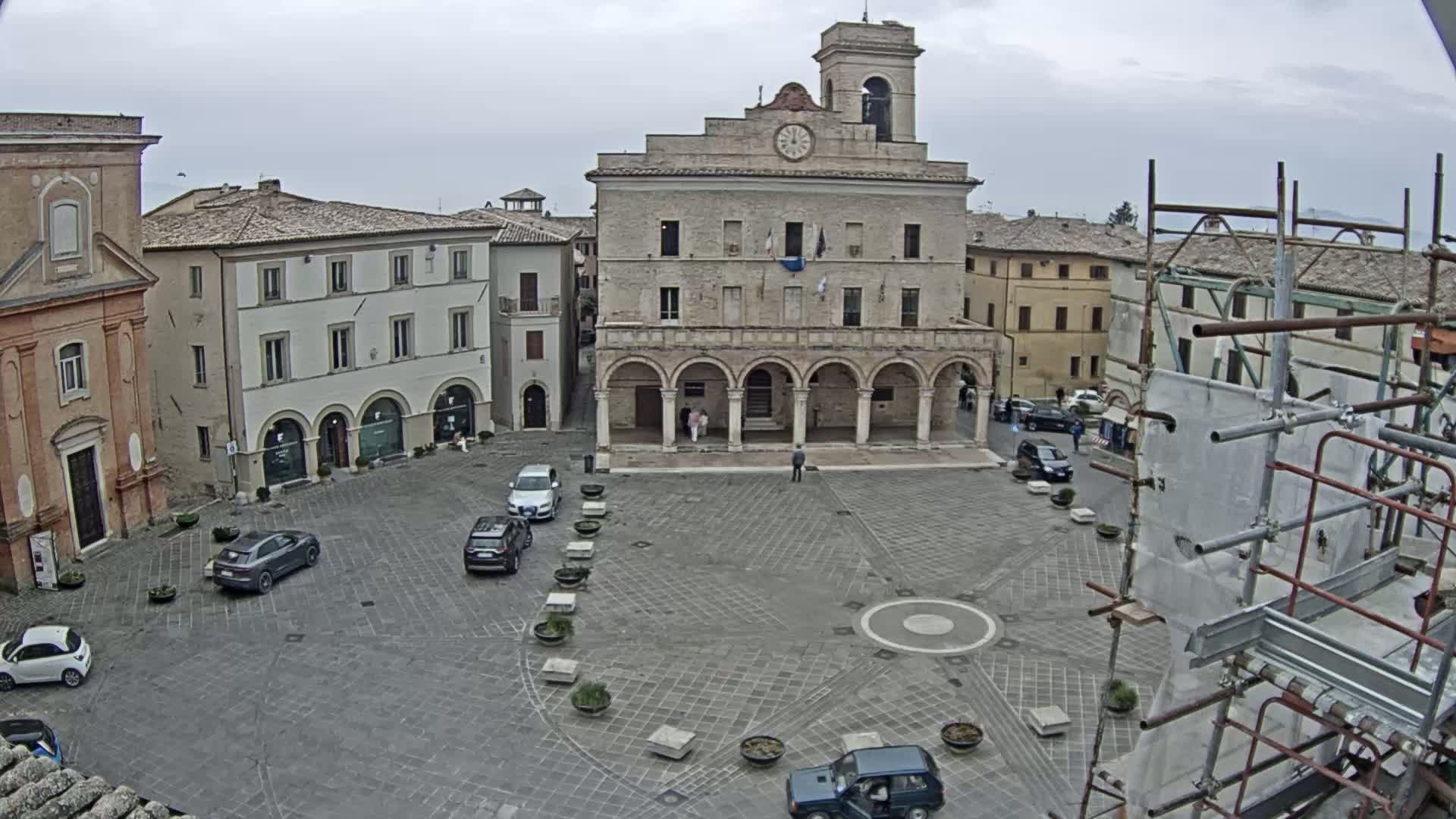 A town square with several cars parked, surrounded by historic buildings under an overcast sky, with scaffolding visible in the bottom right corner.