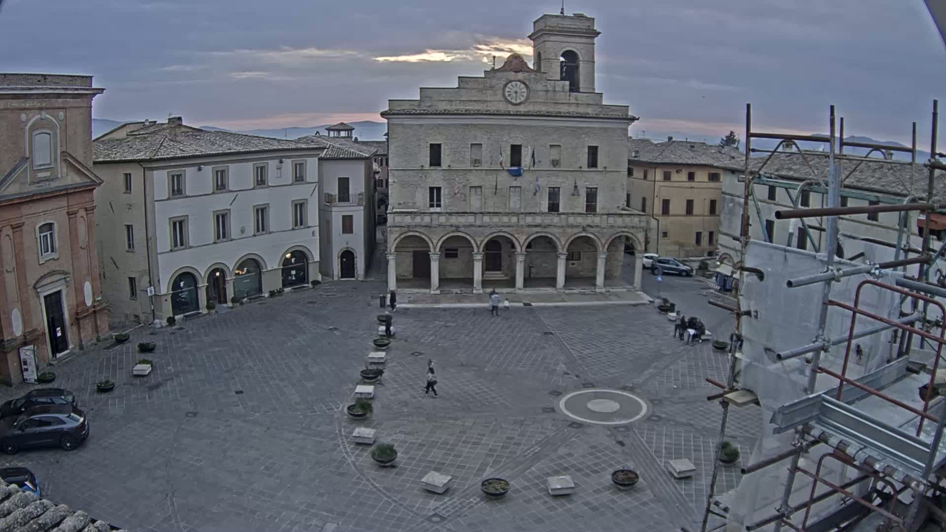A town square with several buildings, including a large stone building with arched entryways, under a cloudy, dusky sky.