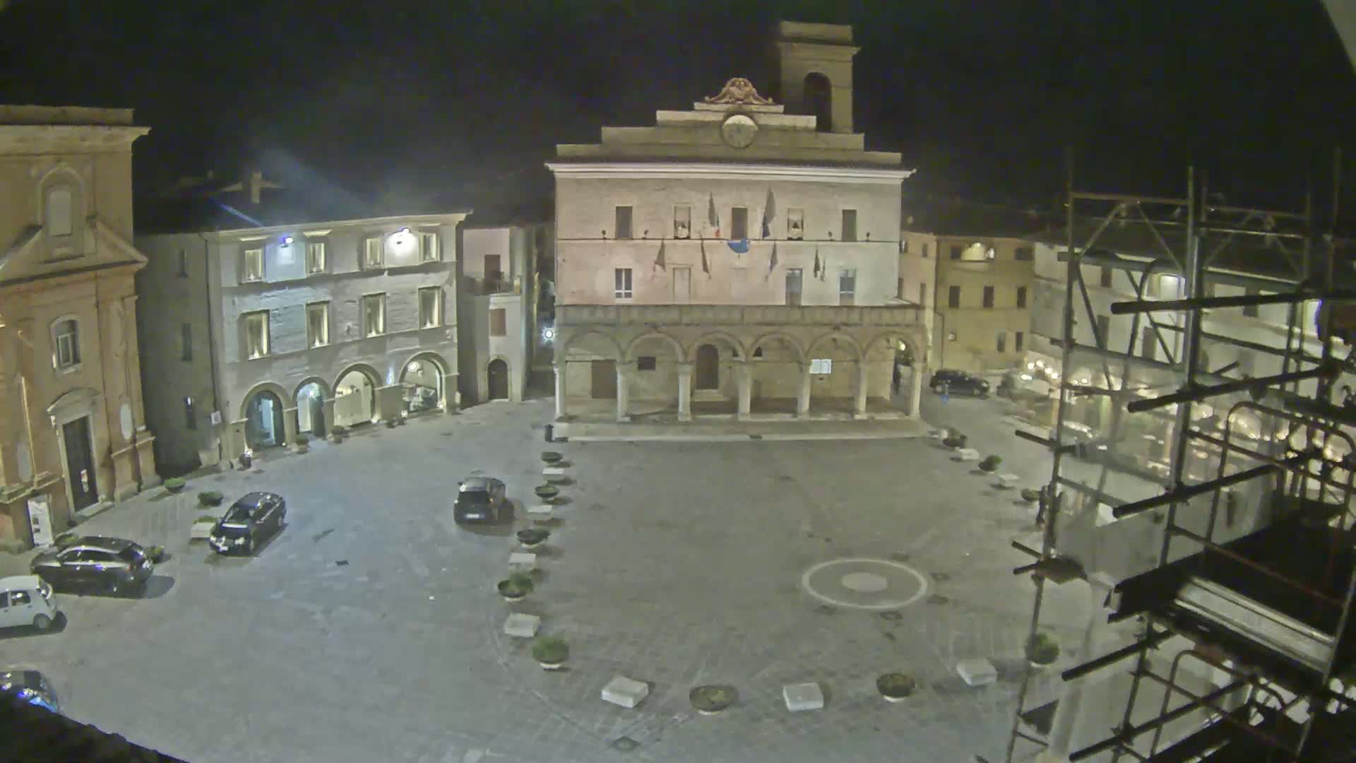 A nighttime view of a town square with several buildings, a few cars parked, and scaffolding surrounding one of the buildings.