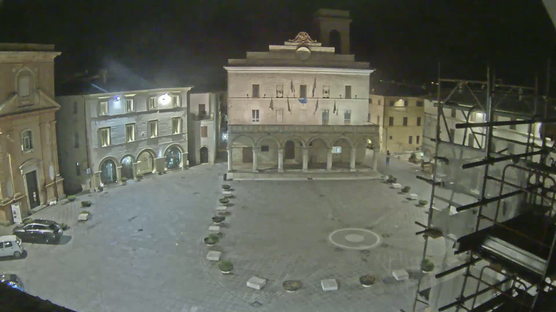 A nighttime view of a town square, surrounded by historic buildings, with scaffolding visible on one side under a dark sky.