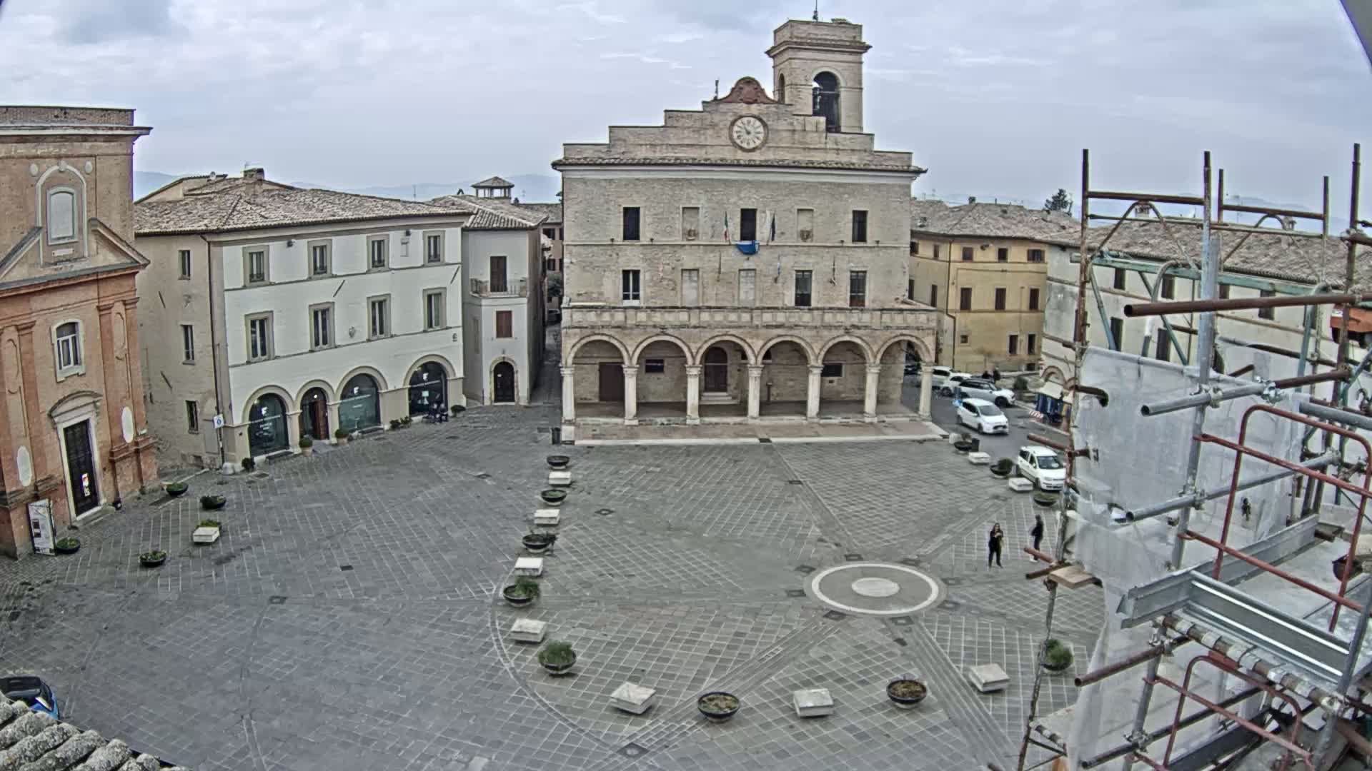 A large town square with a stone building featuring arched entryways and a clock tower, surrounded by other buildings, under an overcast sky.