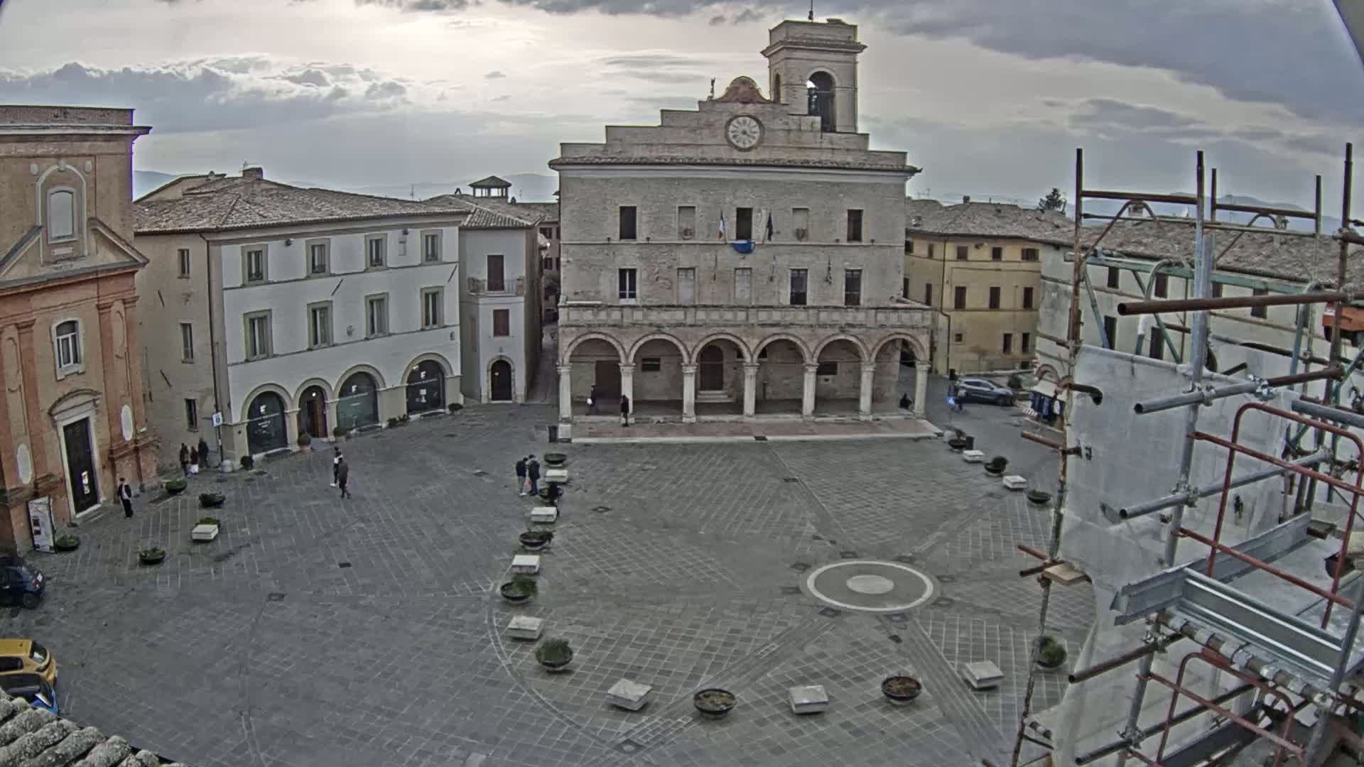 A large town square, under an overcast sky, features a prominent building with arched colonnades and a clock tower, surrounded by other buildings, and a few people are present in the square.