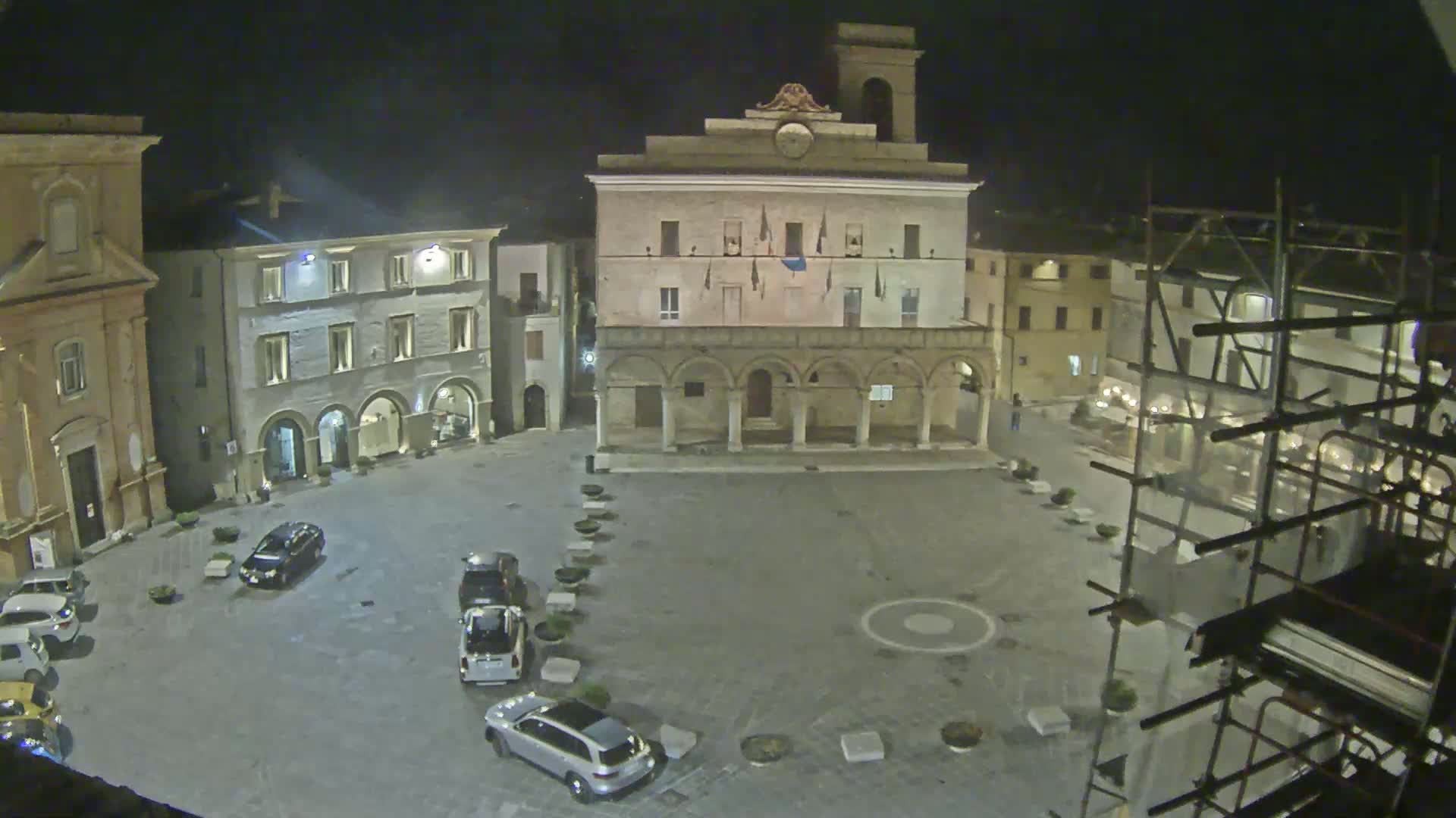 A nighttime view of a town square with several cars parked, surrounded by historic buildings, one of which has scaffolding around it.