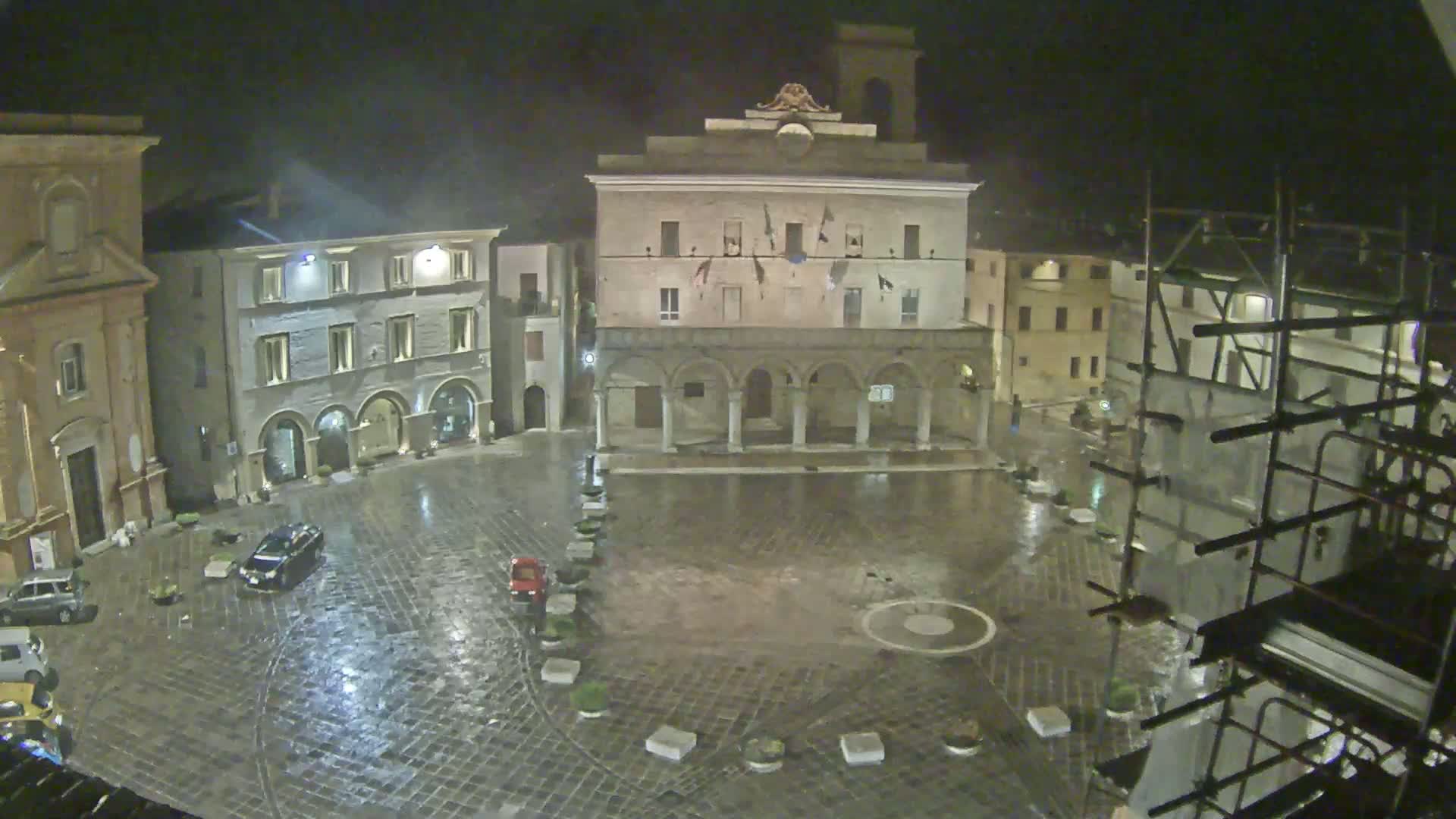 A rain-slicked town square at night, surrounded by stone buildings, one of which has an arcade.