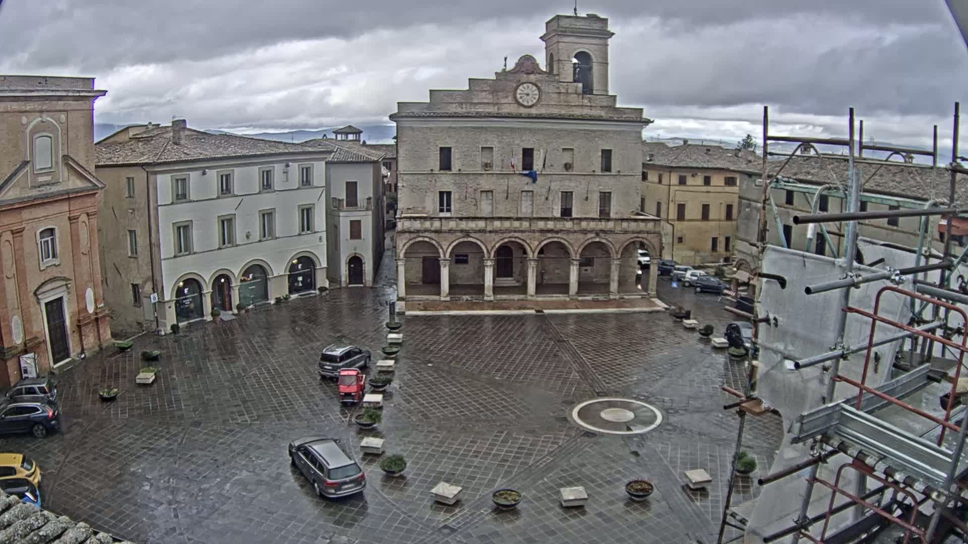 A wet, paved town square is centered on a large building with arched entryways and a clock tower, surrounded by other buildings under a cloudy sky.