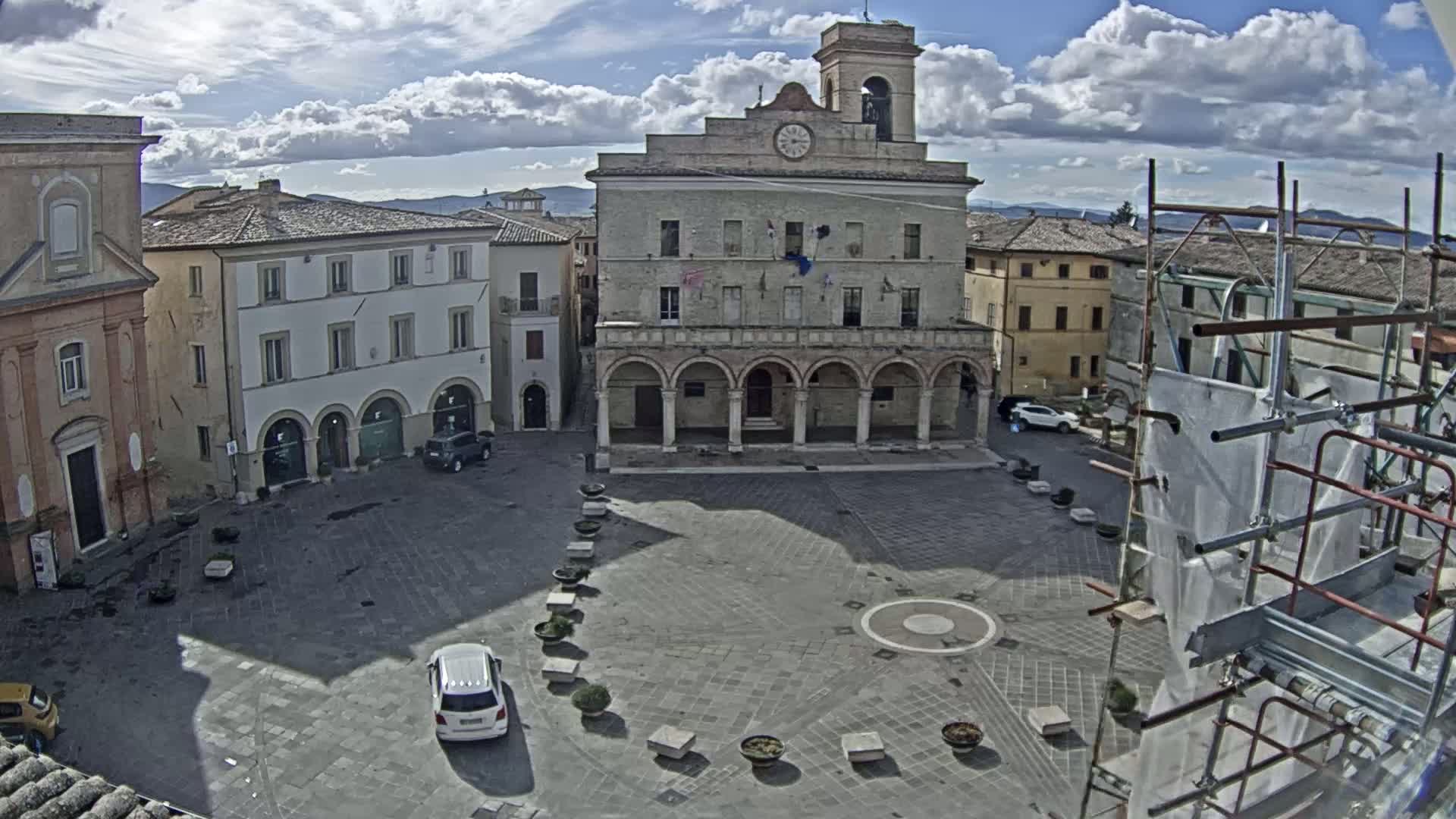 A mostly sunny day shows a town square with a large, arched building, several smaller buildings, and a few cars parked in the square.