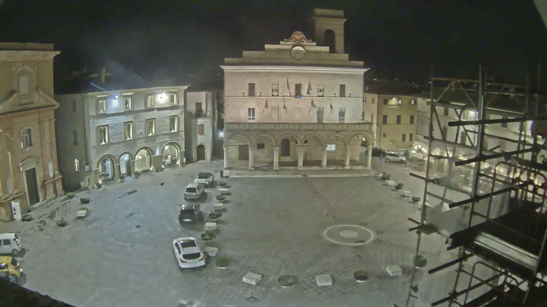 A nighttime view of a town square with several cars parked, surrounded by historic buildings, one of which has scaffolding.
