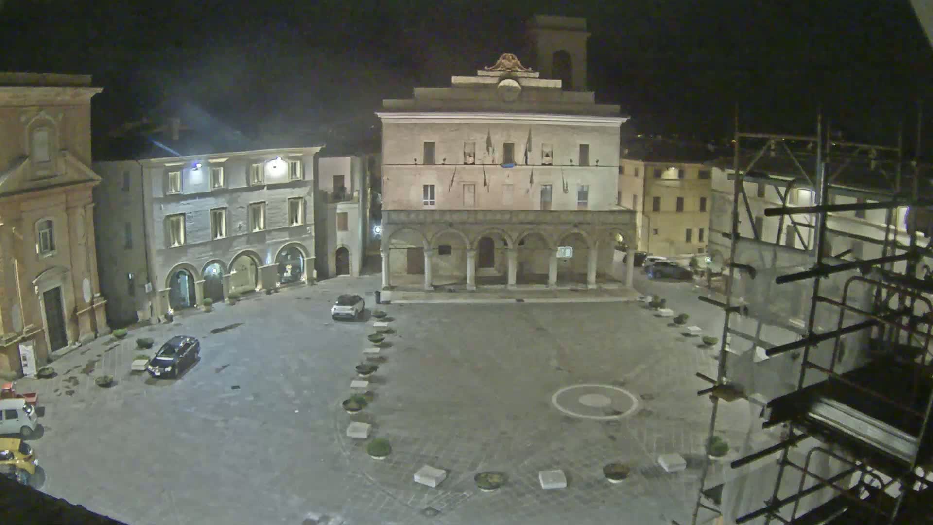 A nighttime view of a town square, featuring several buildings including a large structure with arches, and a few cars parked in the mostly empty square.