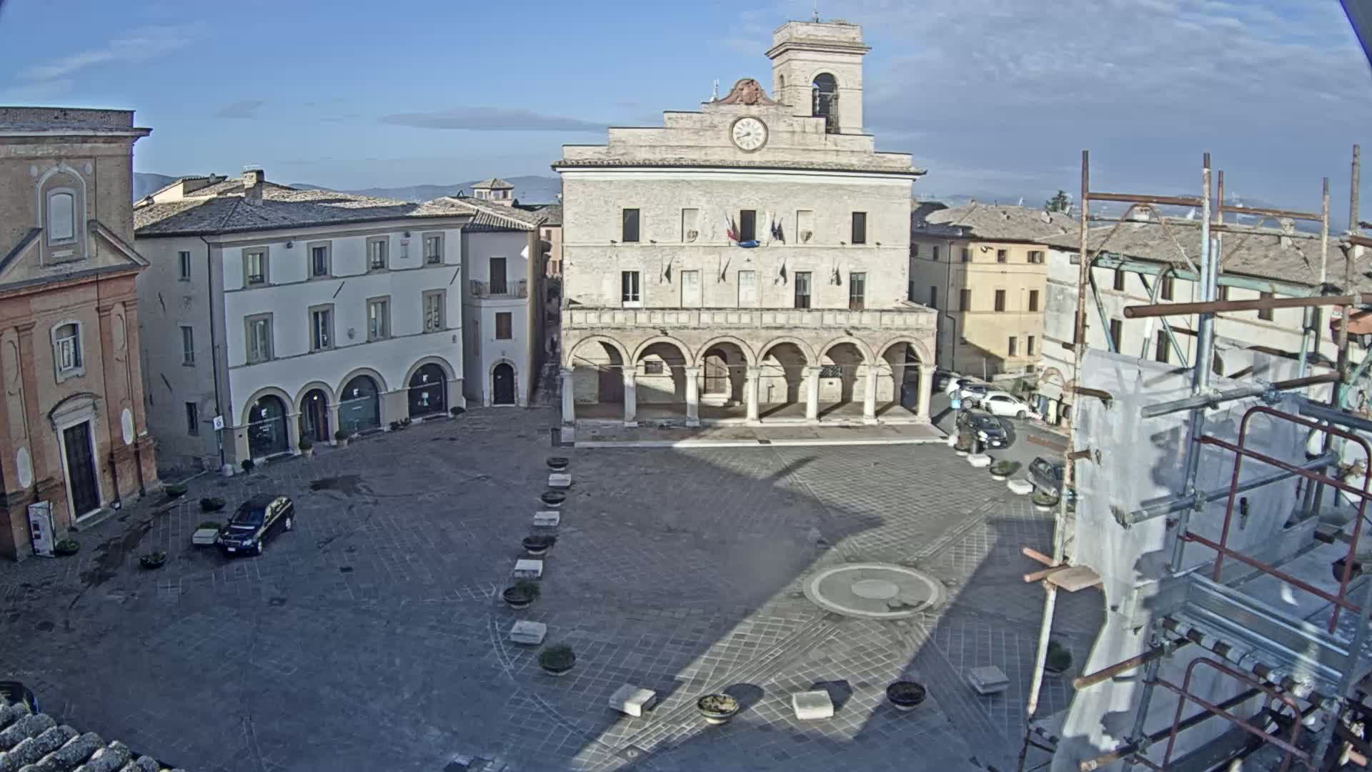 A town square with a large building featuring arched walkways is seen under a clear, sunny sky, with scaffolding visible near the edge of the square.