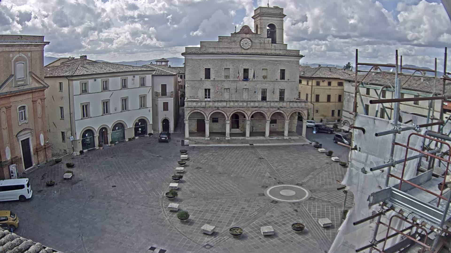 A large, empty town square with a stone building featuring arched entryways and a clock tower is seen under a cloudy sky, with scaffolding visible in the lower right corner.