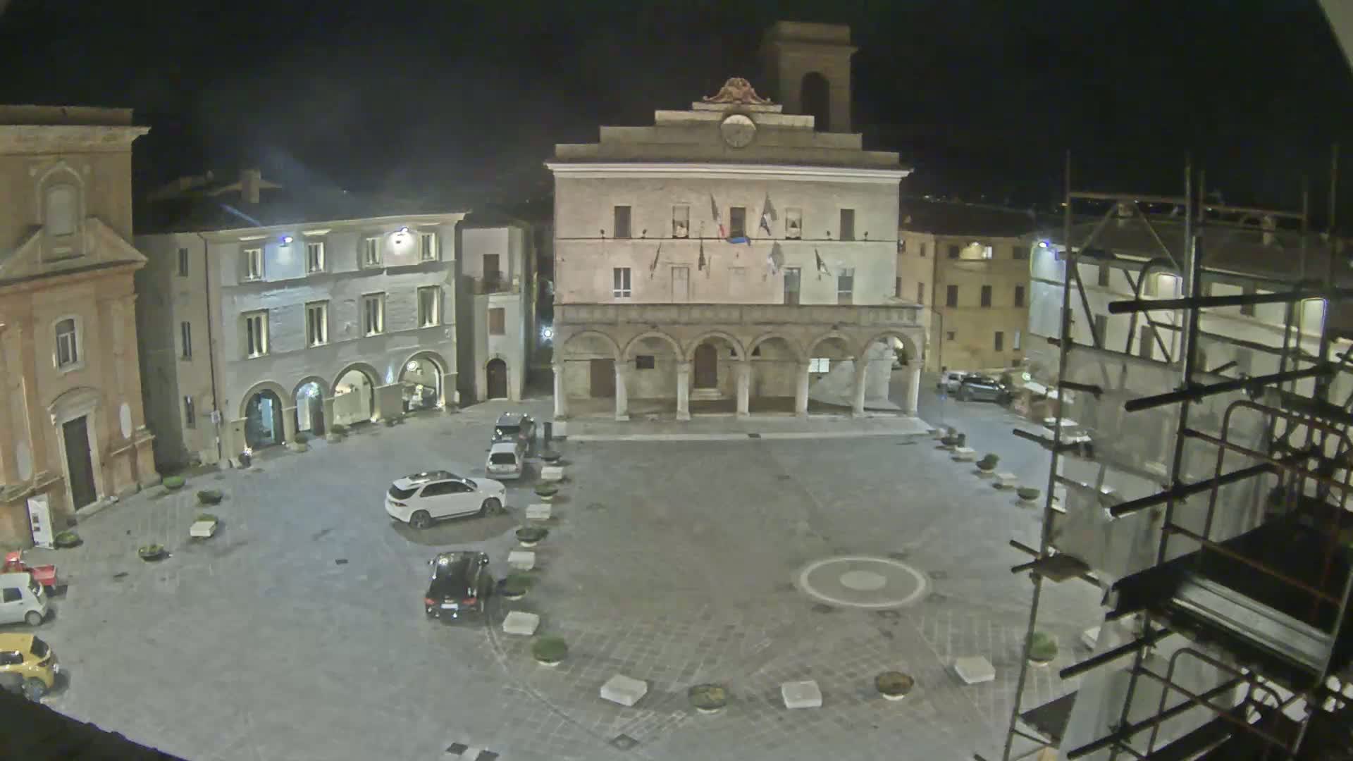 A nighttime view of a town square with several buildings, including a large stone structure with arches, and a few cars parked around the perimeter.