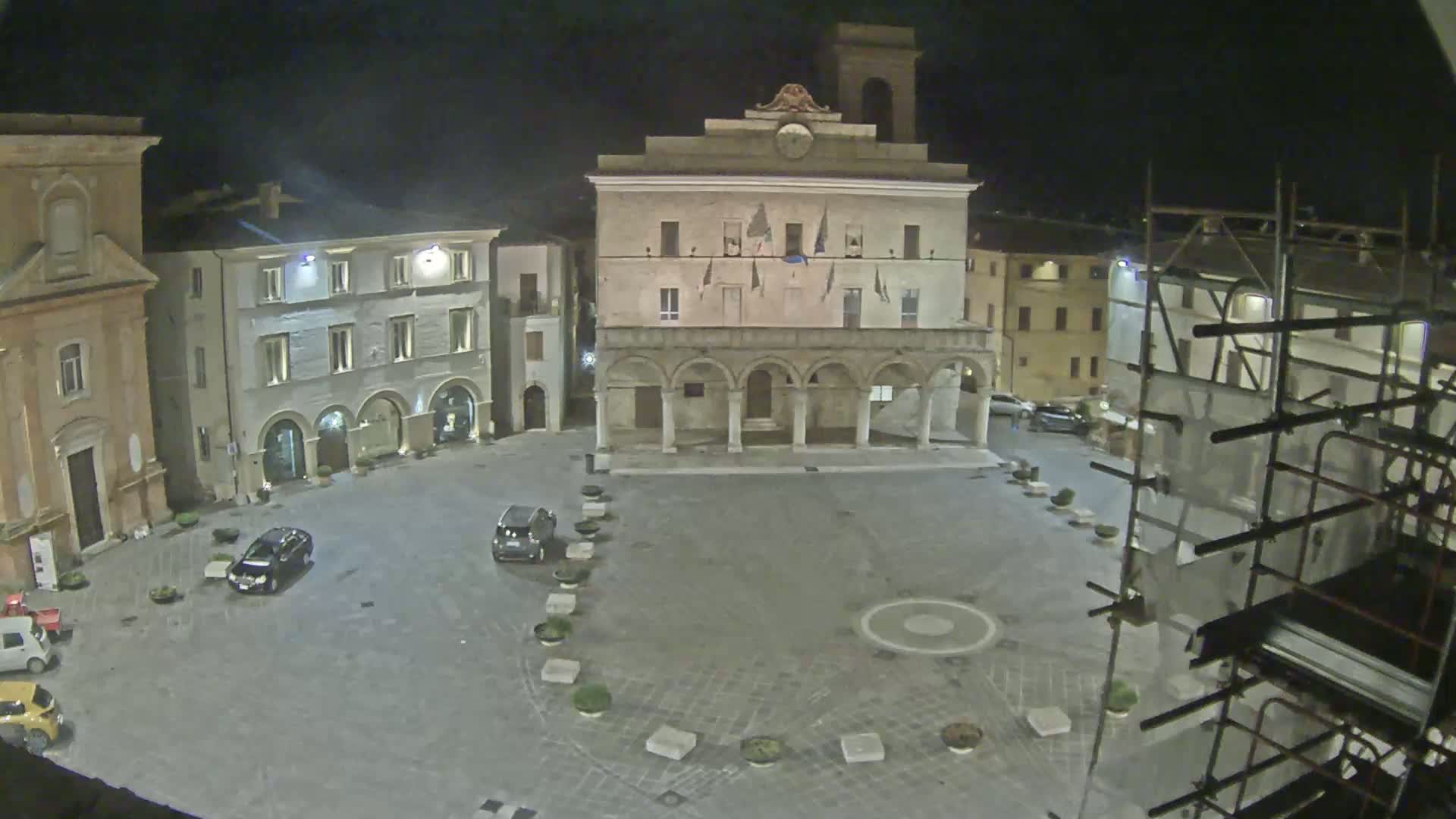 A nighttime view of a town square with several buildings, a few parked cars, and scaffolding on one side of the square.