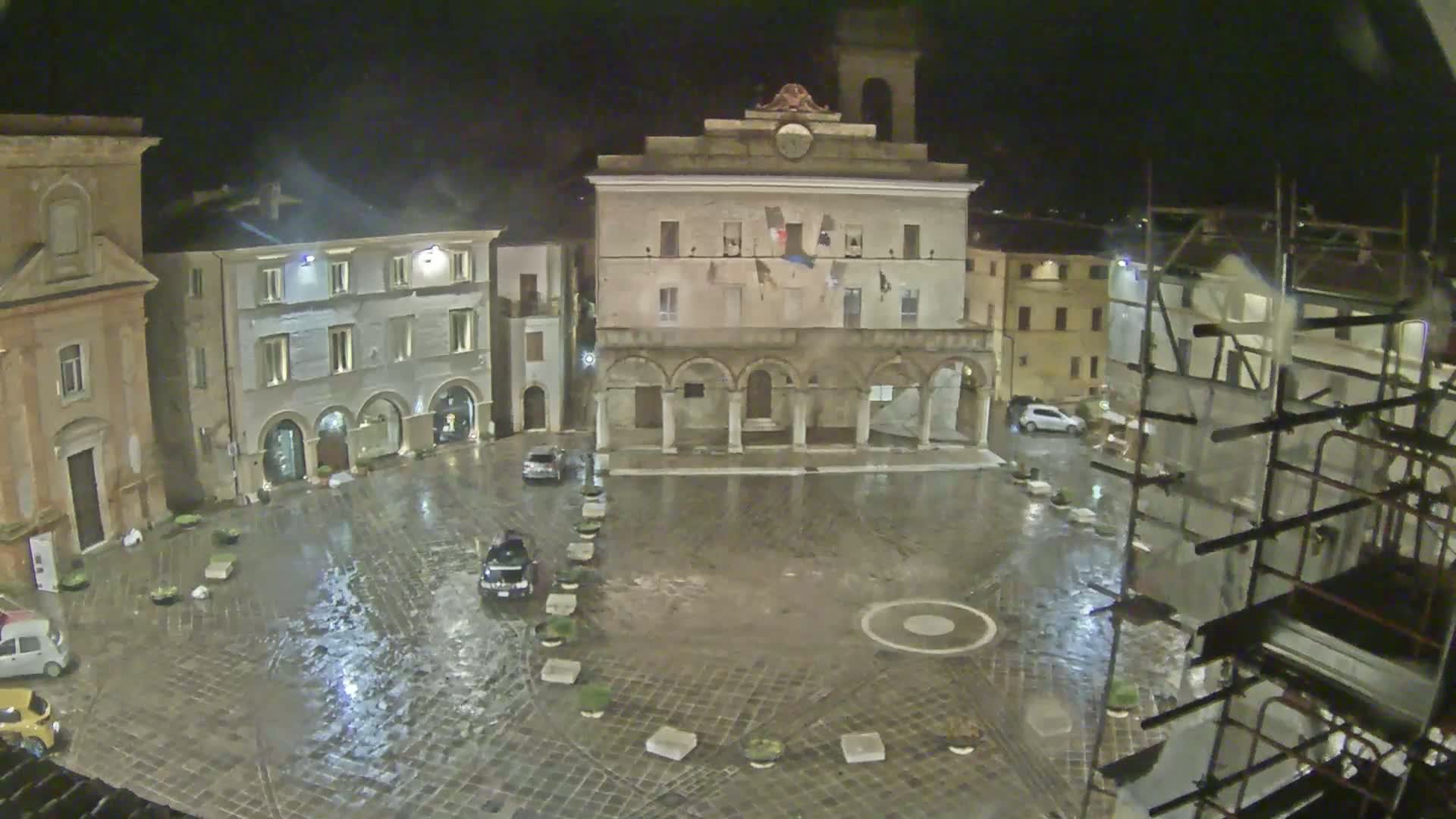 A rain-soaked town square at night, surrounded by historic buildings, with scaffolding visible on one side.