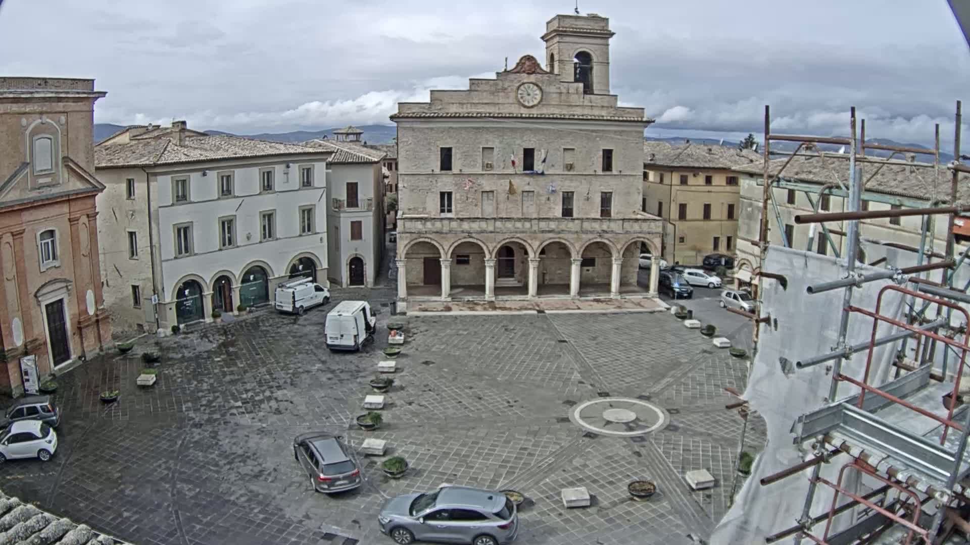 A town square with several cars, historic buildings, and scaffolding under an overcast sky.