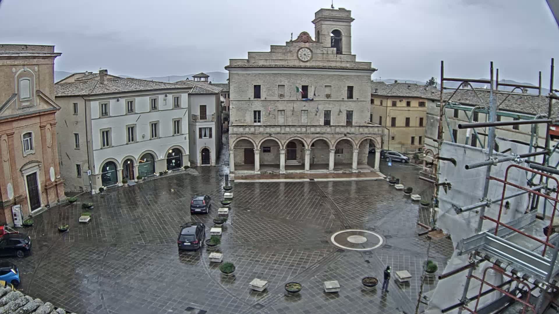 A wet, paved town square is centered on a large stone building with arched entryways, surrounded by other buildings under an overcast sky.