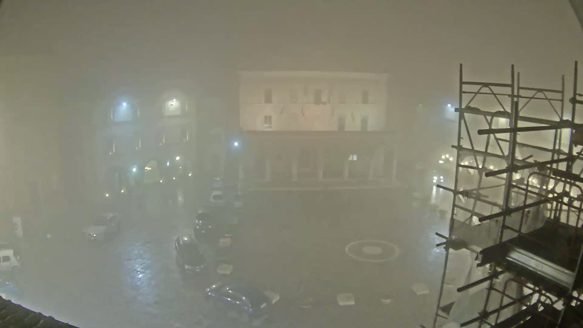 A foggy night scene shows a square with parked cars and a building under construction, all dimly lit.