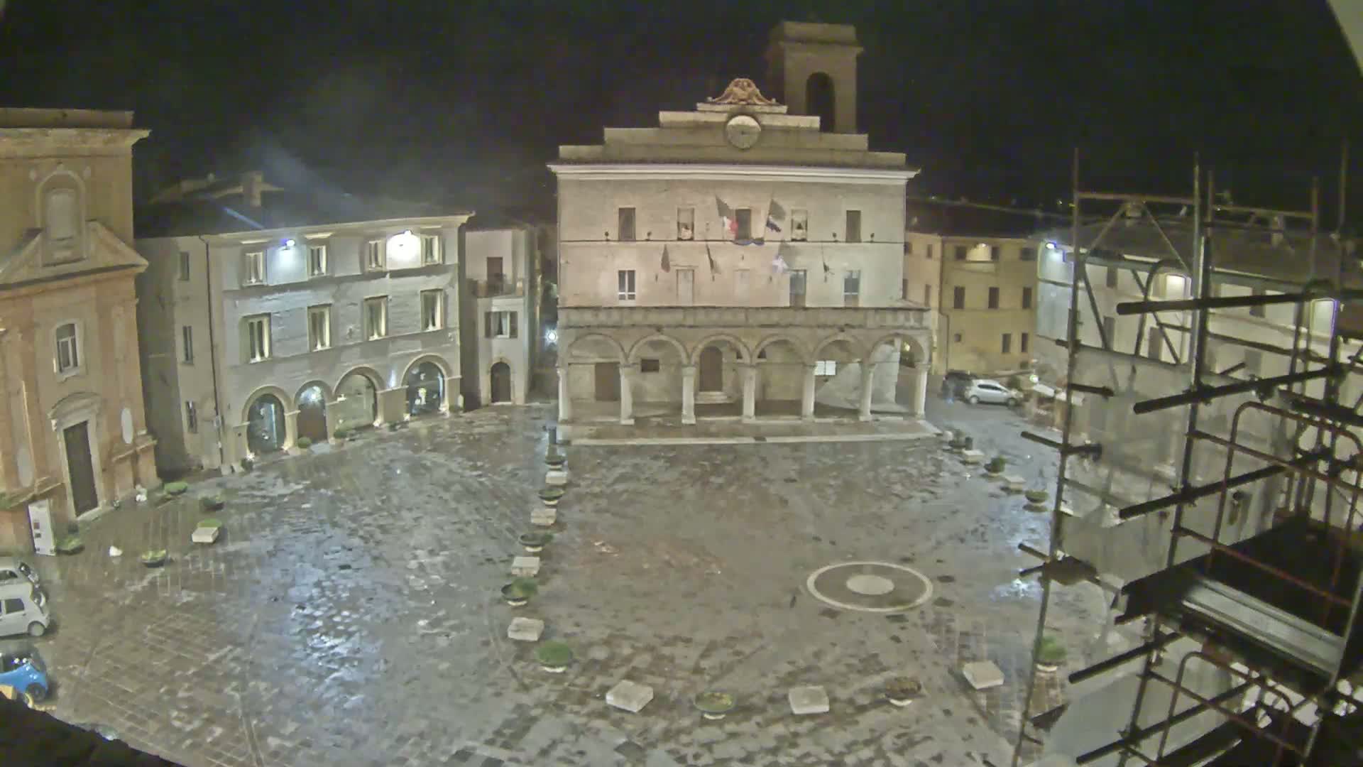 A wet, empty town square at night is surrounded by historic buildings, one of which is under construction.