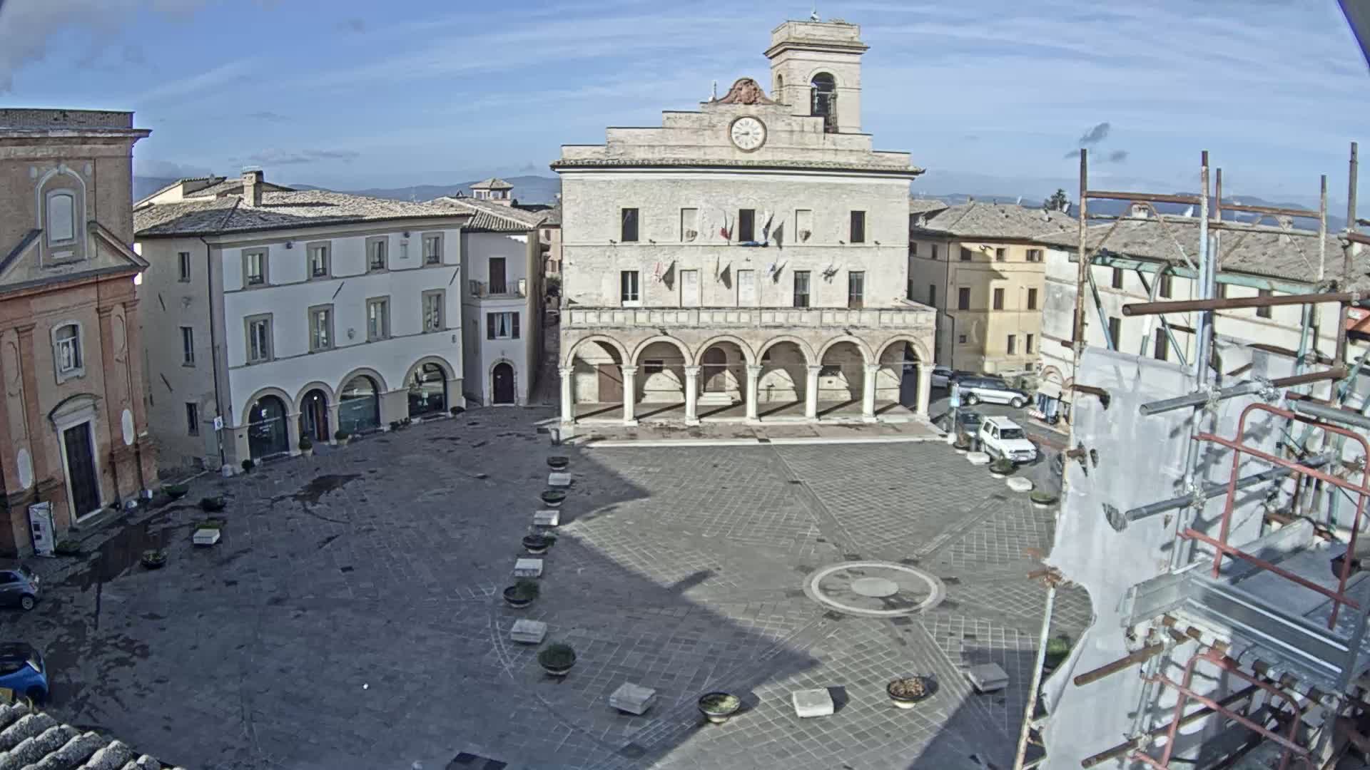 A town square with a large building featuring arches and a clock tower, surrounded by other buildings under a mostly clear sky.