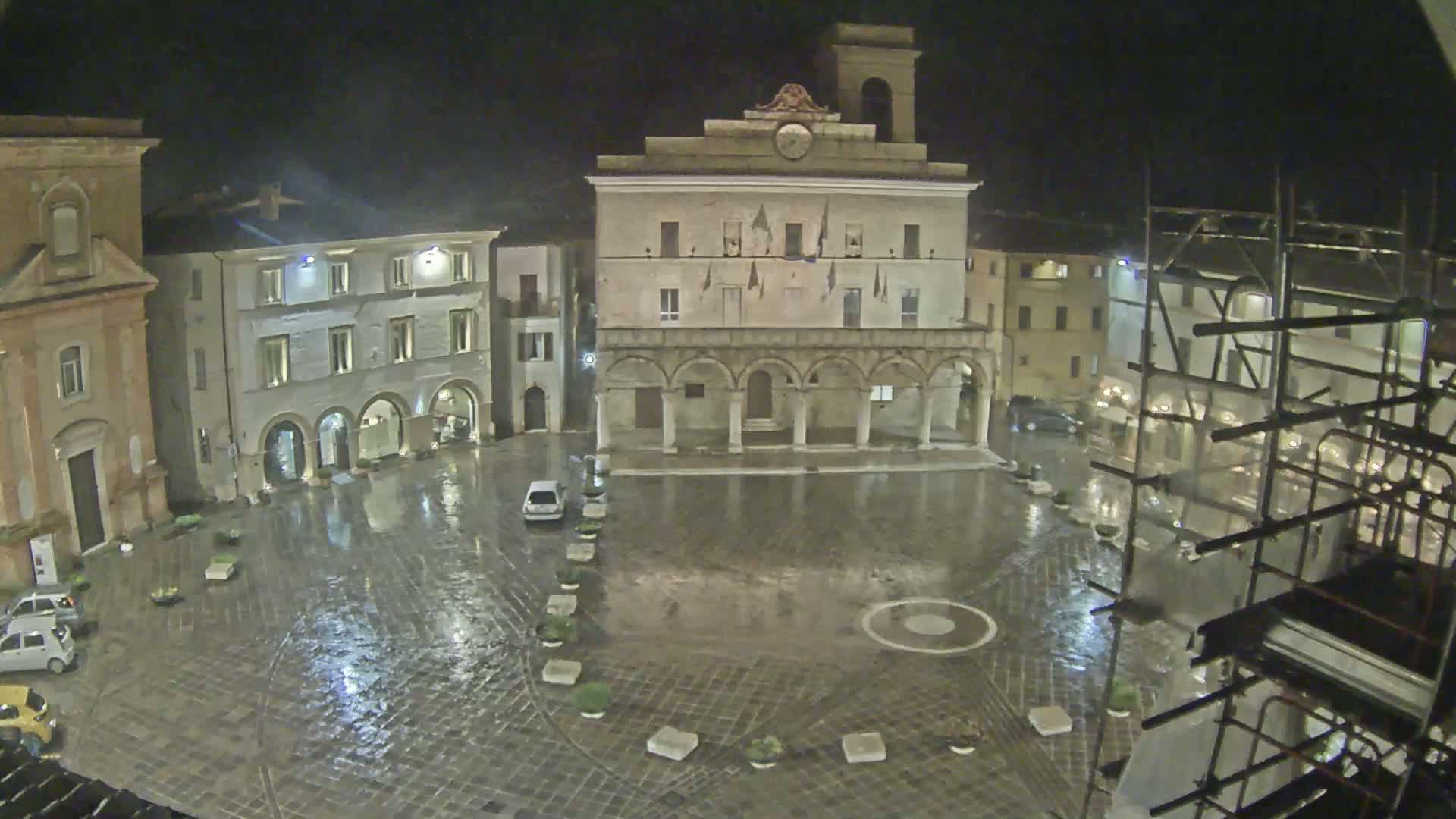 A rain-slicked town square at night, surrounded by historic buildings, one of which is under construction.
