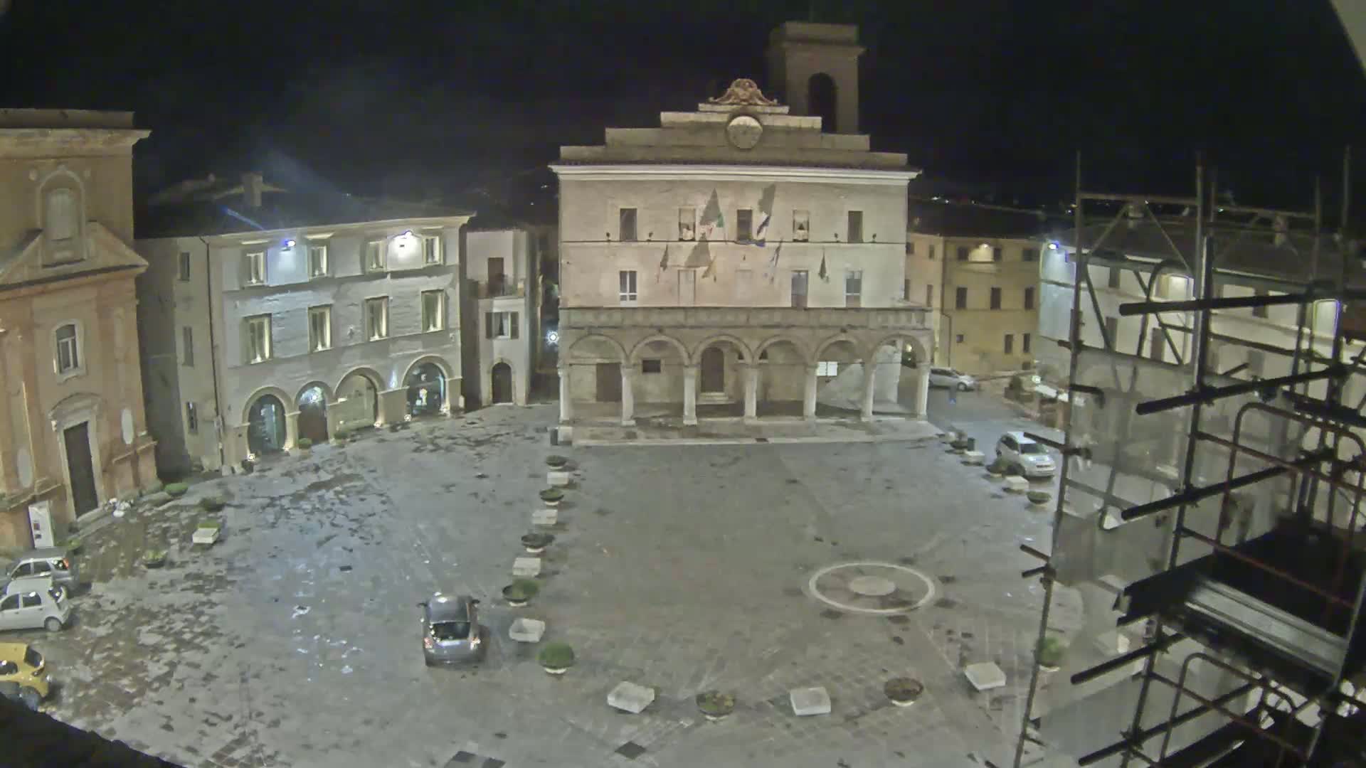 A nighttime view of a town square with several buildings surrounding it, some with scaffolding, and a few cars parked, under wet conditions.