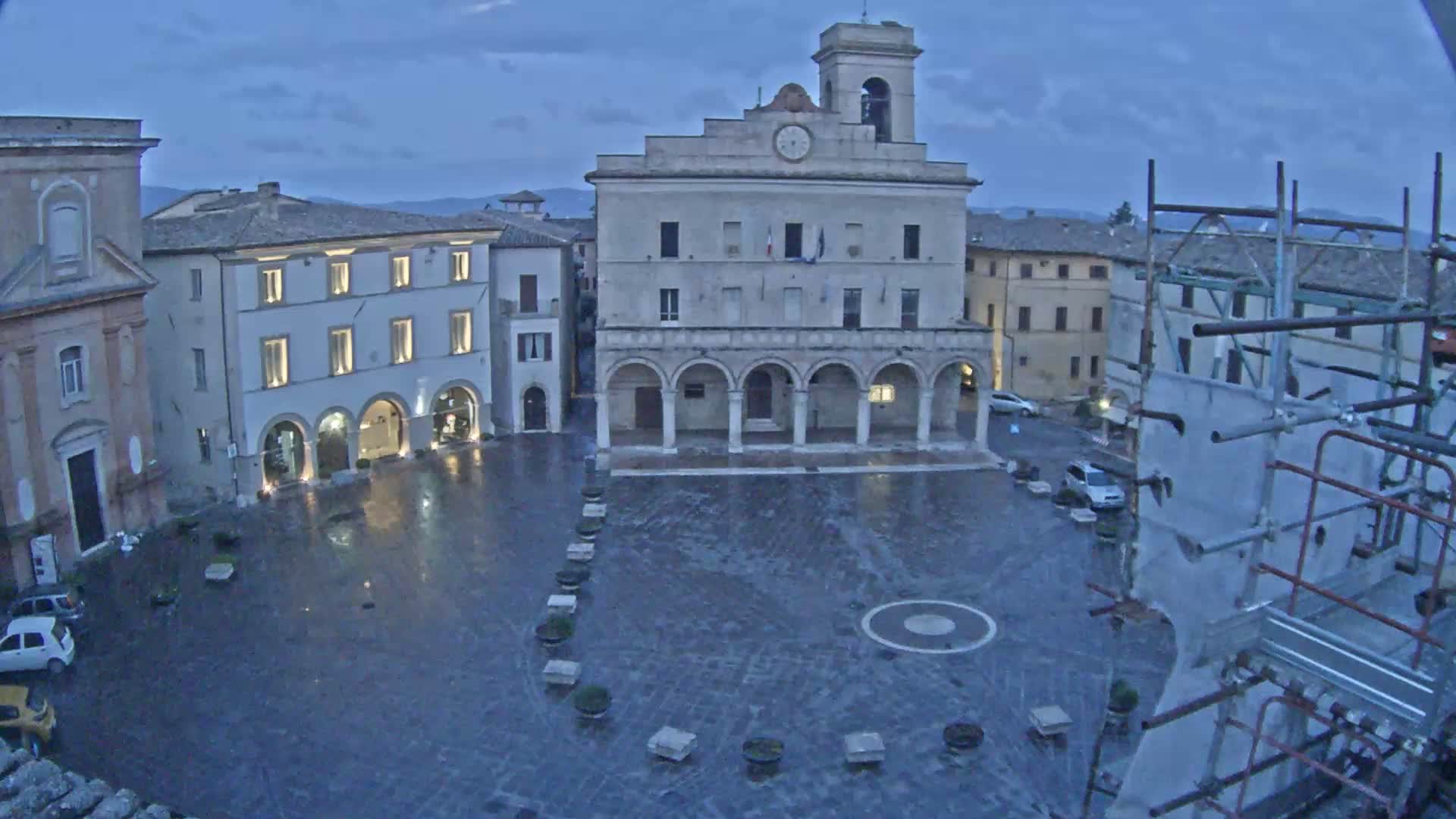 A wet, stone plaza is surrounded by historic buildings under a twilight sky, with scaffolding visible in the foreground.