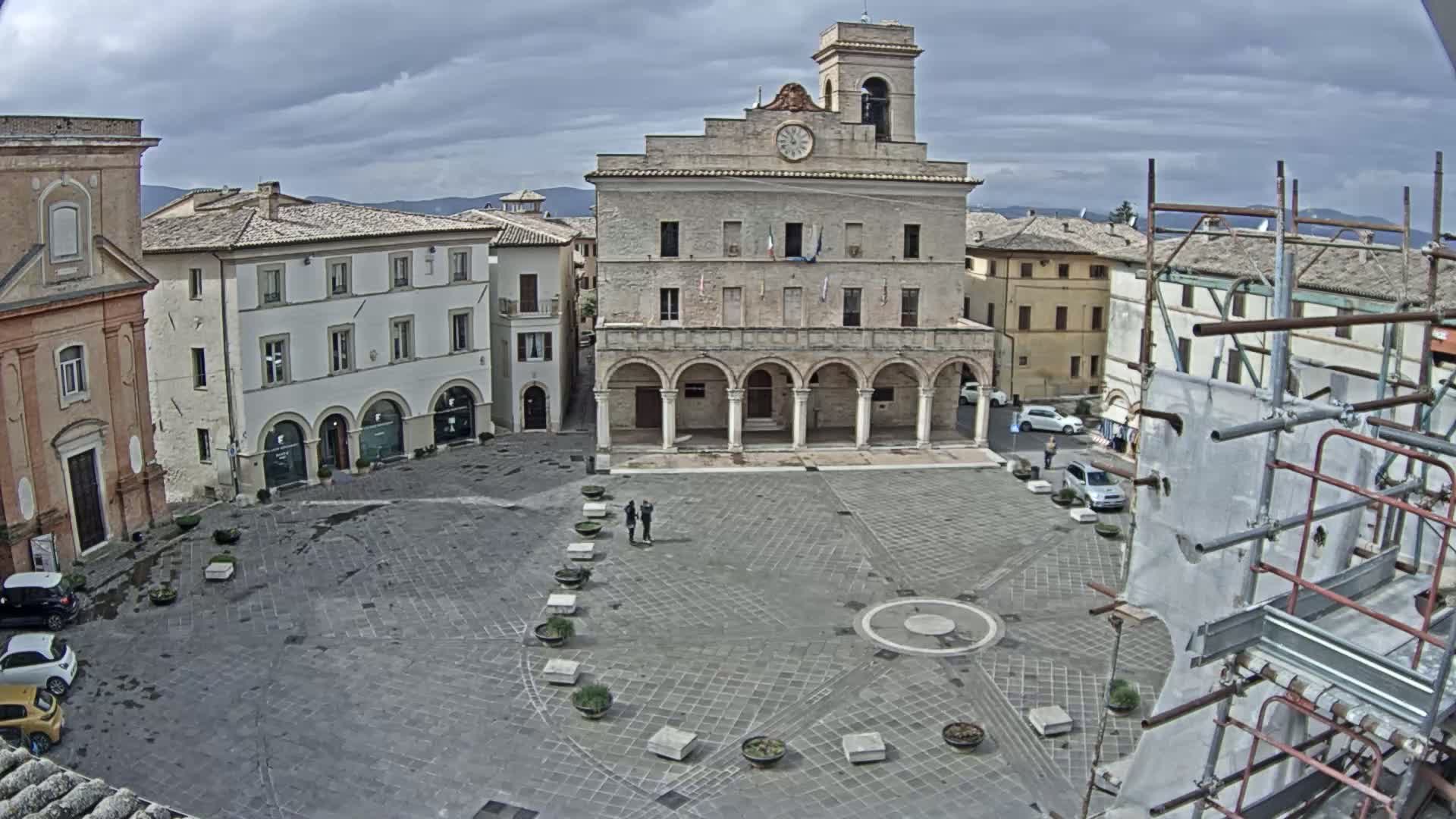 A town square with a large stone building, several smaller buildings, and a few cars under a cloudy sky.
