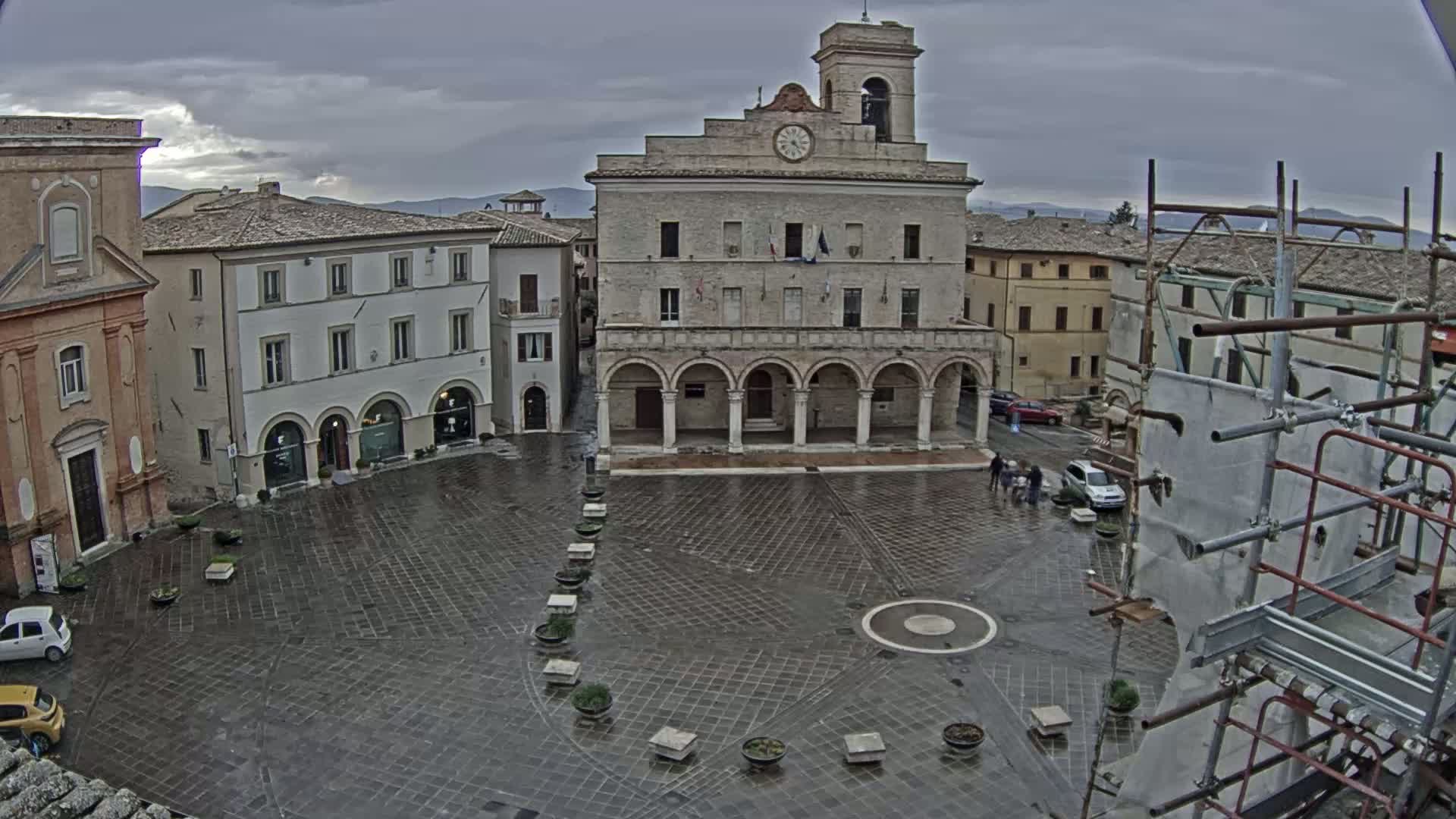 A town square with a large stone building and clock tower is seen on an overcast day, with a few people and cars present, and scaffolding to the right.