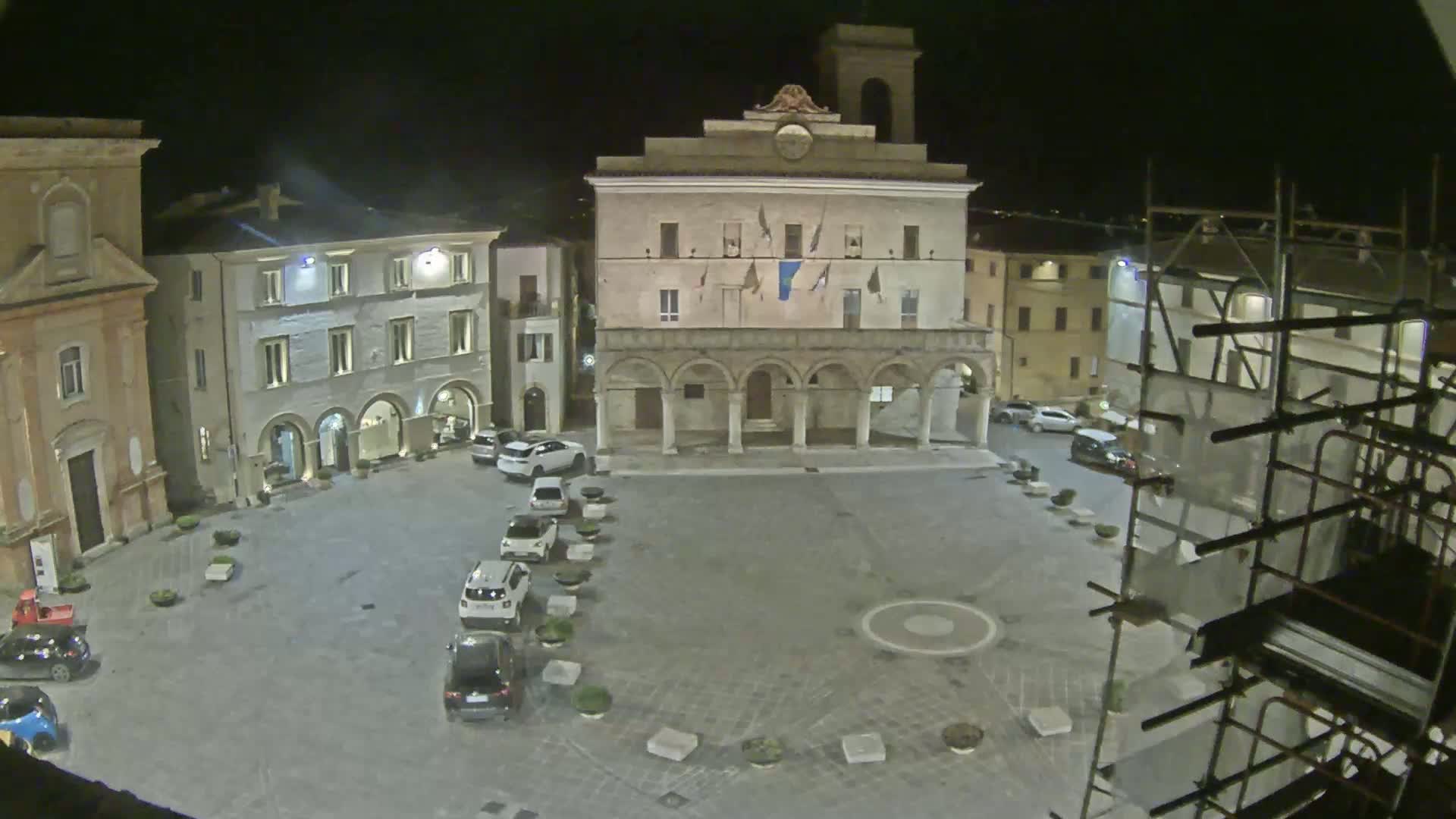 A nighttime view of a town square with several cars parked along one side, surrounded by buildings, one of which has scaffolding.