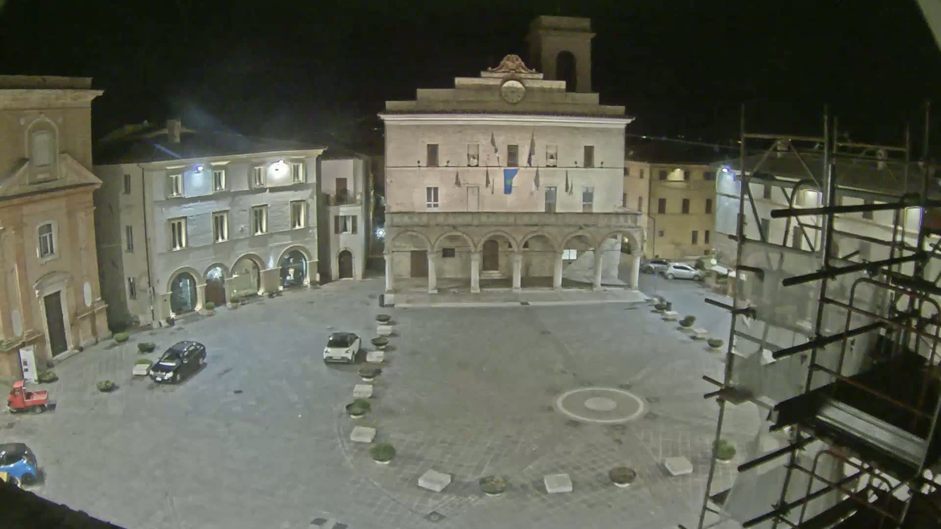 A nighttime view of a town square with several buildings, including a large, arched building at the center, a few cars parked, and scaffolding on the right.